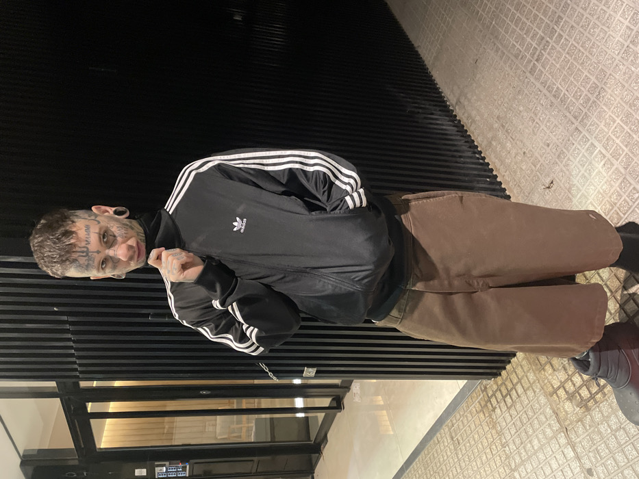A single individual stands centrally on an urban sidewalk, illuminated at night, in front of a modern building in Buenos Aires, Argentina. The person, appearing male, faces slightly right of the camera with a direct gaze. He possesses short, dark, textured hair and extensive facial tattoos, including text on the temples ("MAMMI" visible on the right), black stud piercings on the nose bridge and above the right eyebrow, and large black ear gauges. He is dressed in a black turtleneck worn under a black Adidas track jacket featuring white stripes on the sleeves and a white Adidas logo on the left chest. Below, he wears brown, wide-leg, knee-length shorts and dark grey/black insulated boot-like footwear over dark socks. His right hand grasps the collar of the turtleneck, while his left hand rests inside the pocket of the track jacket. The background to the left displays a glass building entrance door with a visible interior and a hanging metal chain. To the right, a dark wall with vertical ribbed paneling is present. The sidewalk is paved with light grey square-patterned tiles.
