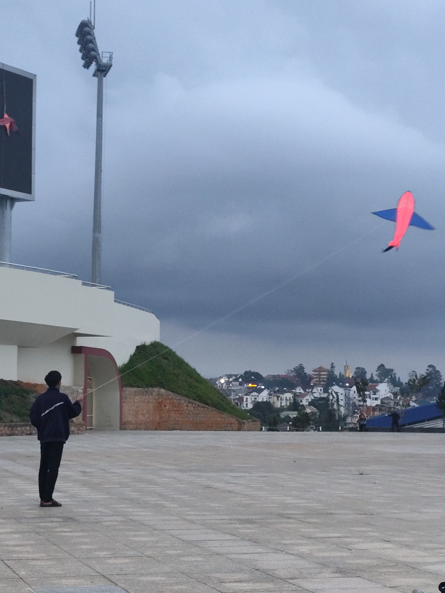 The image captures a person flying a kite in the city of Lang Biang, Da Lat, Vietnam. The kite, shaped like an airplane, is predominantly red with blue wings and tail, and is visible in the upper right quadrant of the frame. The person, seen from behind and in profile, is standing in a paved outdoor area, wearing a dark jacket and dark pants, and holding the kite string. In the background, a white building with a curved structure and green grassy slopes are visible, suggesting a park or a stadium complex. Further in the distance, a town with various buildings, some with tiled roofs, is nestled among trees, and a cloudy, overcast sky completes the scene. A tall light pole with a bank of stadium lights is present on the left side of the image.