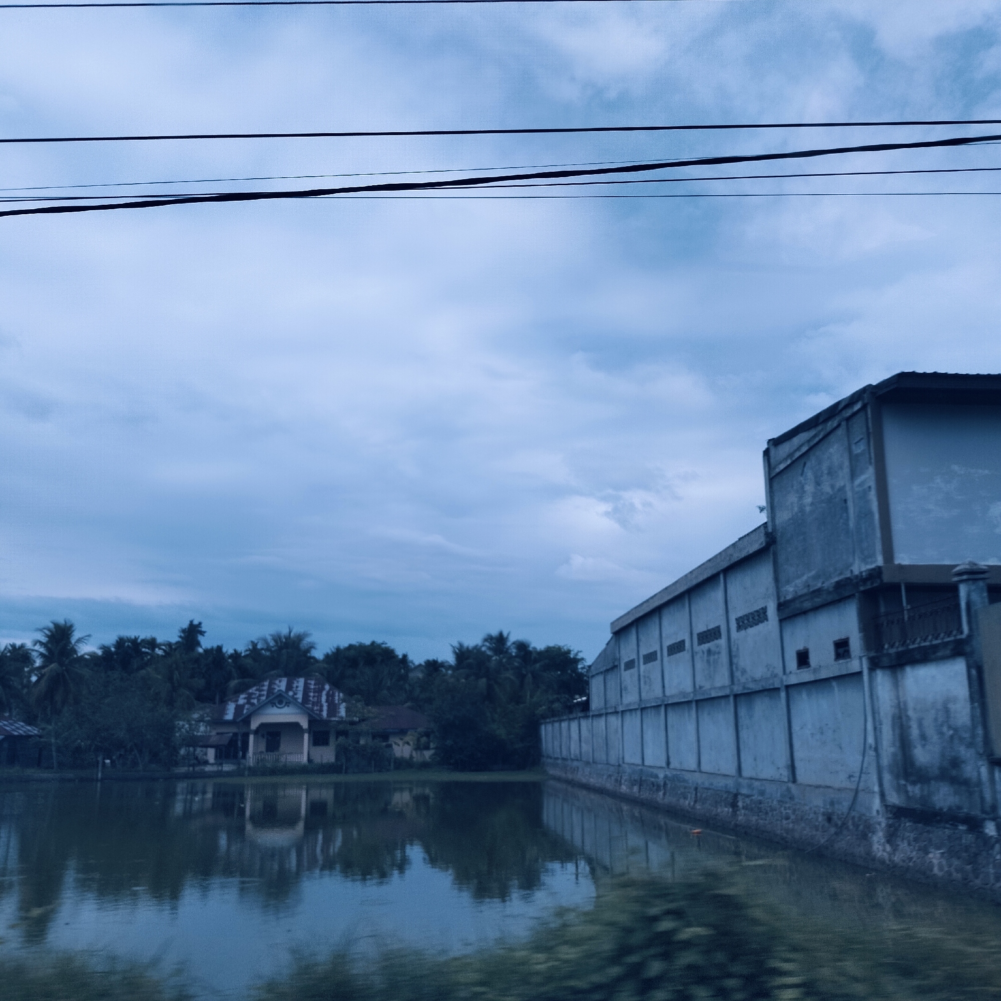 The image captures a quiet, overcast landscape in what appears to be Payameuneng, Indonesia, featuring a still body of water, buildings, and dense tropical foliage. The upper third of the frame is dominated by a light blue-grey sky with soft, diffused clouds, suggesting an overcast day or perhaps early morning/late afternoon. Several dark, horizontal power lines traverse the sky, slightly sagging.
A tranquil expanse of water occupies the mid-ground, serving as a mirror to the sky and the structures along its banks. On the right, a large, grey, industrial-looking concrete building with a flat roof stretches along the water's edge. Its long side features multiple regularly spaced, rectangular openings or vents. The base of the wall appears to be constructed from a rougher material, possibly stone or textured concrete, and a section with dark railings is visible towards the right end of the building. The structure's reflection is clearly cast upon the calm water.
To the left, across the water and nestled among lush greenery, stands a residential house. It features a distinctive, possibly corrugated roof and a small porch. The house itself appears to be a light, faded color, perhaps cream or pale yellow, with subtle decorative details on its facade. Dense tropical vegetation, including numerous palm trees, forms a verdant backdrop to the house and extends throughout the background, characteristic of the region. The immediate foreground shows blurry green foliage, possibly indicating the photograph was taken from a moving perspective.
No people are visible in the scene, and there is no discernible text. The overall atmosphere is one of stillness and the blending of human construction with the abundant natural environment under a muted sky.