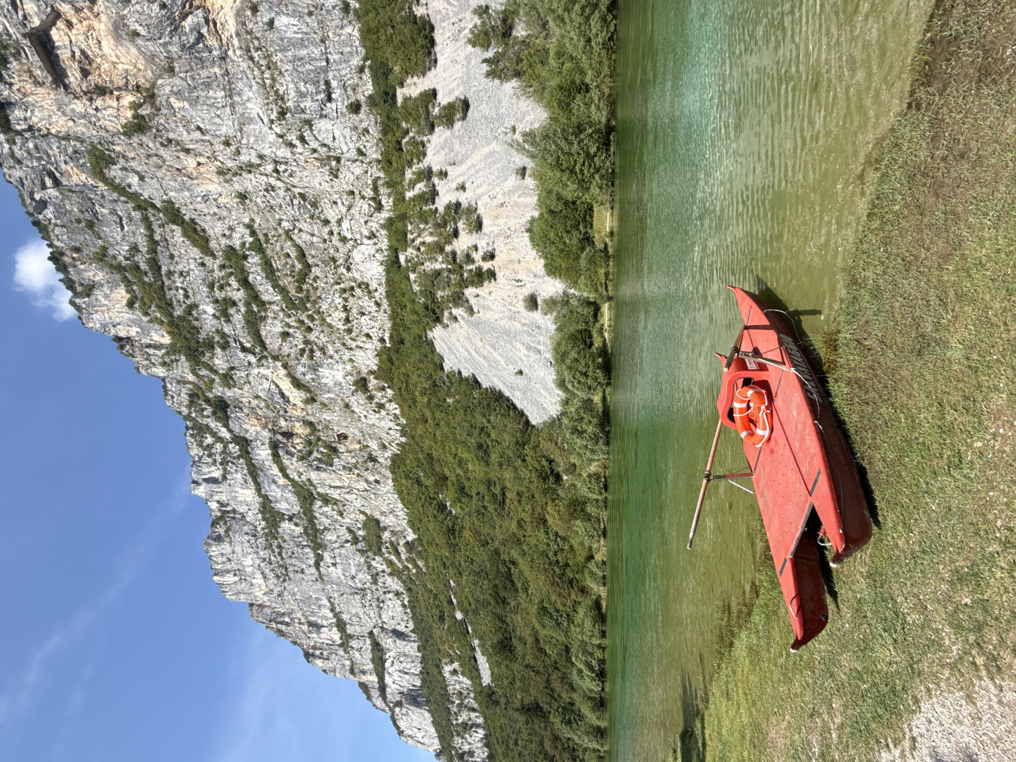 A bright red, two-hulled boat with a life preserver rests on the grassy bank of a clear, green lake. The boat is positioned in the lower left of the frame. In the background, a massive, sheer grey mountain face rises dramatically, partially covered by dense green trees that cling to its slopes. The sky above is a clear, vibrant blue with a few wispy clouds. The scene depicts a serene natural landscape.
