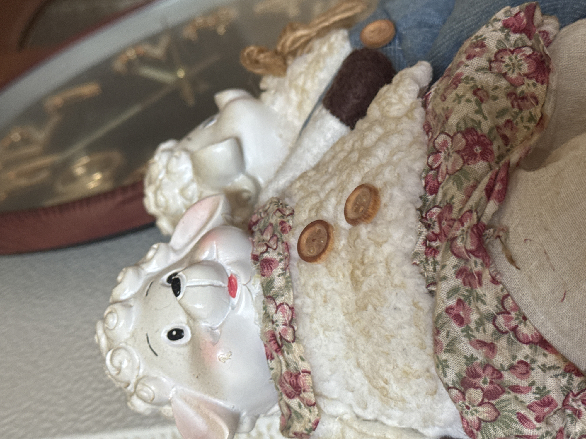 The image displays two decorative sheep figurines. The primary figurine in the foreground is white with curly wool-like texture on its head and a fluffy white body. It wears a floral-patterned fabric collar with pink and green floral designs, and two round brown buttons on its chest. Its face has black eyes, pink cheeks, and a small red mouth. The second figurine, partially visible behind the first, appears similar in construction. The scene is indoors, with a blurred background that includes reflective surfaces and hints of other objects. There is no information within the image to suggest a location in Kholmsk, Russia.