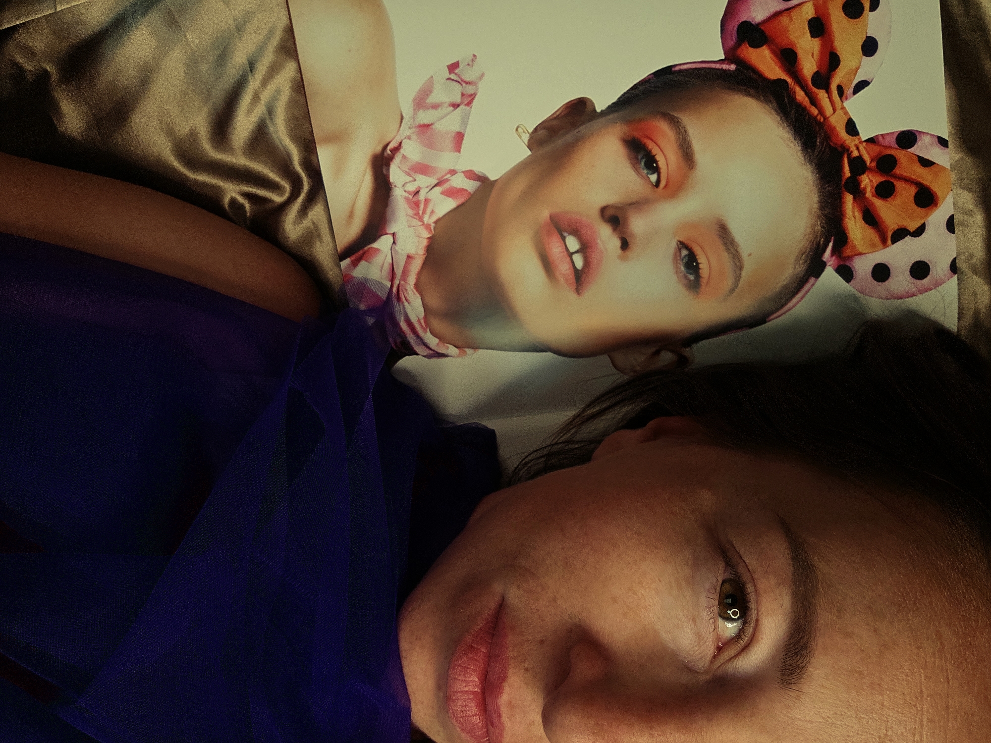 A person is taking a selfie, with a magazine open in the foreground. The magazine features a model wearing a pink and orange bow with black polka dots, orange eyeshadow, and a pink and white striped scarf. The person in the selfie is partially visible, with their eye and lips in focus, and a dark purple fabric draped over their shoulder. The background appears to be a wall. This image was taken in Chaikovsky, Russia.