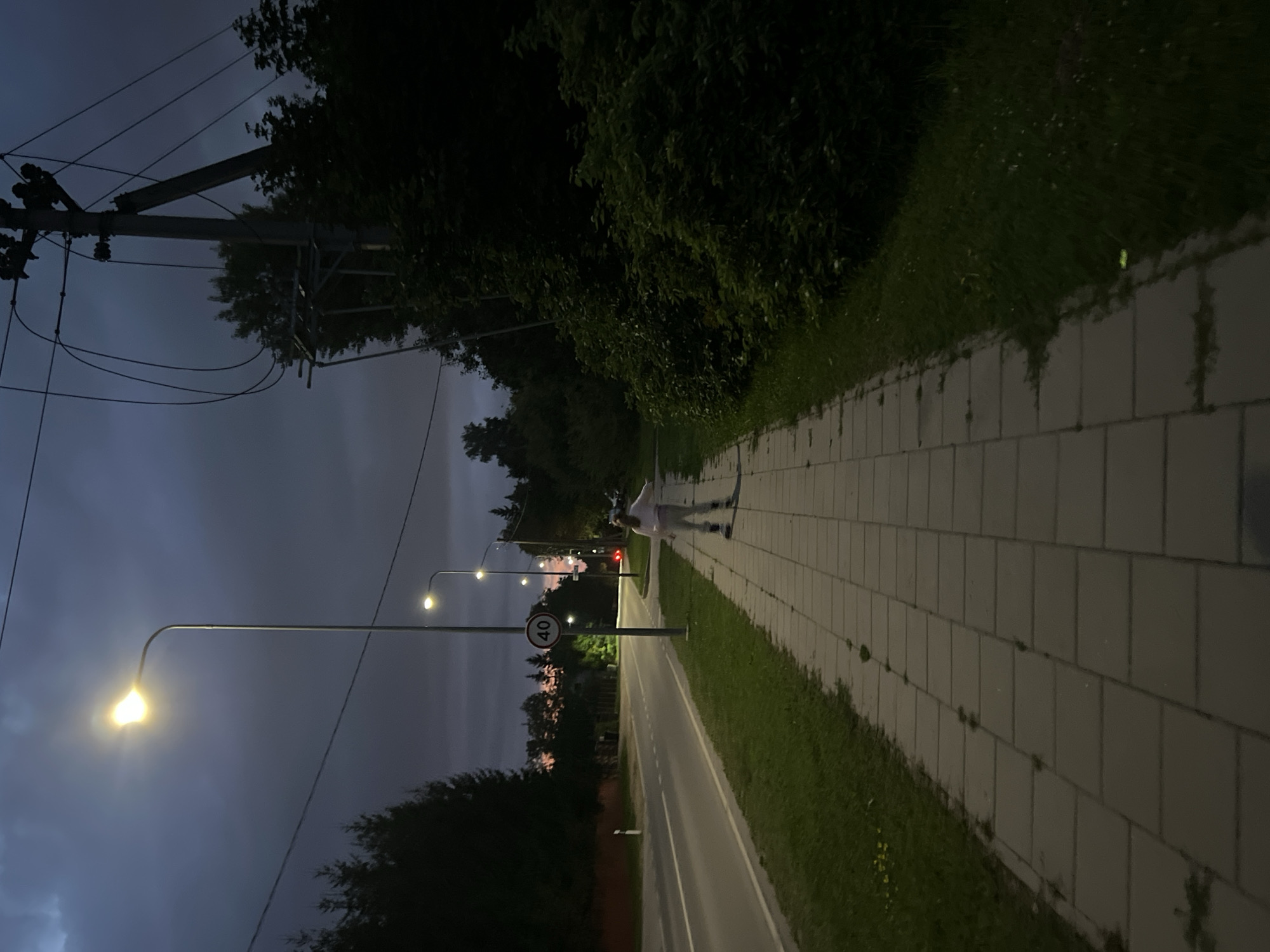 An exterior shot captures a person rollerblading on a paved sidewalk at dusk, adjacent to a road. The person, wearing a light-colored tracksuit, is seen in mid-stride, with their arms extended. A streetlamp illuminates the path. In the background, a speed limit sign with "40" is visible on a pole, and further down the road, a red taillight indicates another vehicle. Trees line the right side of the scene, and a power pole with multiple wires is prominent. The sky is dark with clouds, suggesting evening. The road appears to be a two-lane asphalt surface. The location is likely a suburban or semi-rural area within Vilnius, Lithuania, given the style of streetlights and the presence of power lines and a speed limit sign.