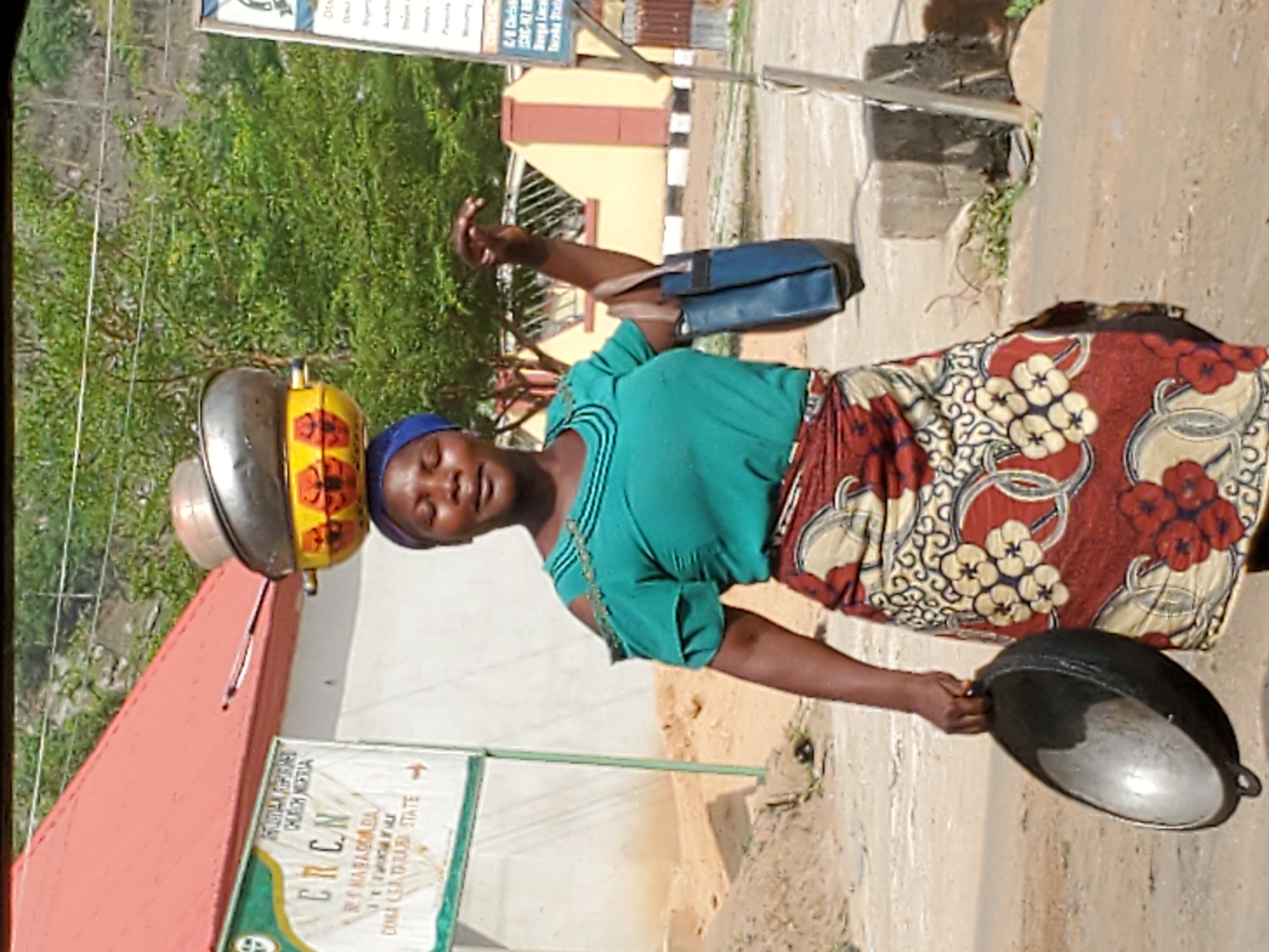 A woman is captured walking outdoors, expertly balancing a stack of cooking pots or bowls on her head. She wears a blue headwrap, a teal-green short-sleeved top, and a vibrant red, white, and yellow patterned skirt (wrapper). On her head, a large silver pot forms the base, topped by a yellow and orange patterned pot, and a smaller silver bowl. A dark blue handbag is slung over her right shoulder, and she holds a large, dark, shallow pan in her left hand.
The setting appears to be a street or pathway with dusty ground. In the background, there are buildings of simple construction; one is yellow with a brown roof section, and another, partly visible on the left, has a red roof. Lush green trees and vegetation are visible behind the buildings.
The image is taken during the daytime, under bright sunlight. The woman looks towards the camera with a calm, slightly smiling expression. A prominent signpost in the background features rotated text, which includes "CRCN", "RMABADABA", and "DOUNA LEA TARASI STATE", providing location context consistent with Mararaba, Taraba State, Nigeria. Additional fragmented text from the sign visible through OCR includes "CHONTHIA", "DAC-NO", "Berga Laca", and "Iscoña Steta". The woman's activity suggests she is likely a vendor or transporting goods.