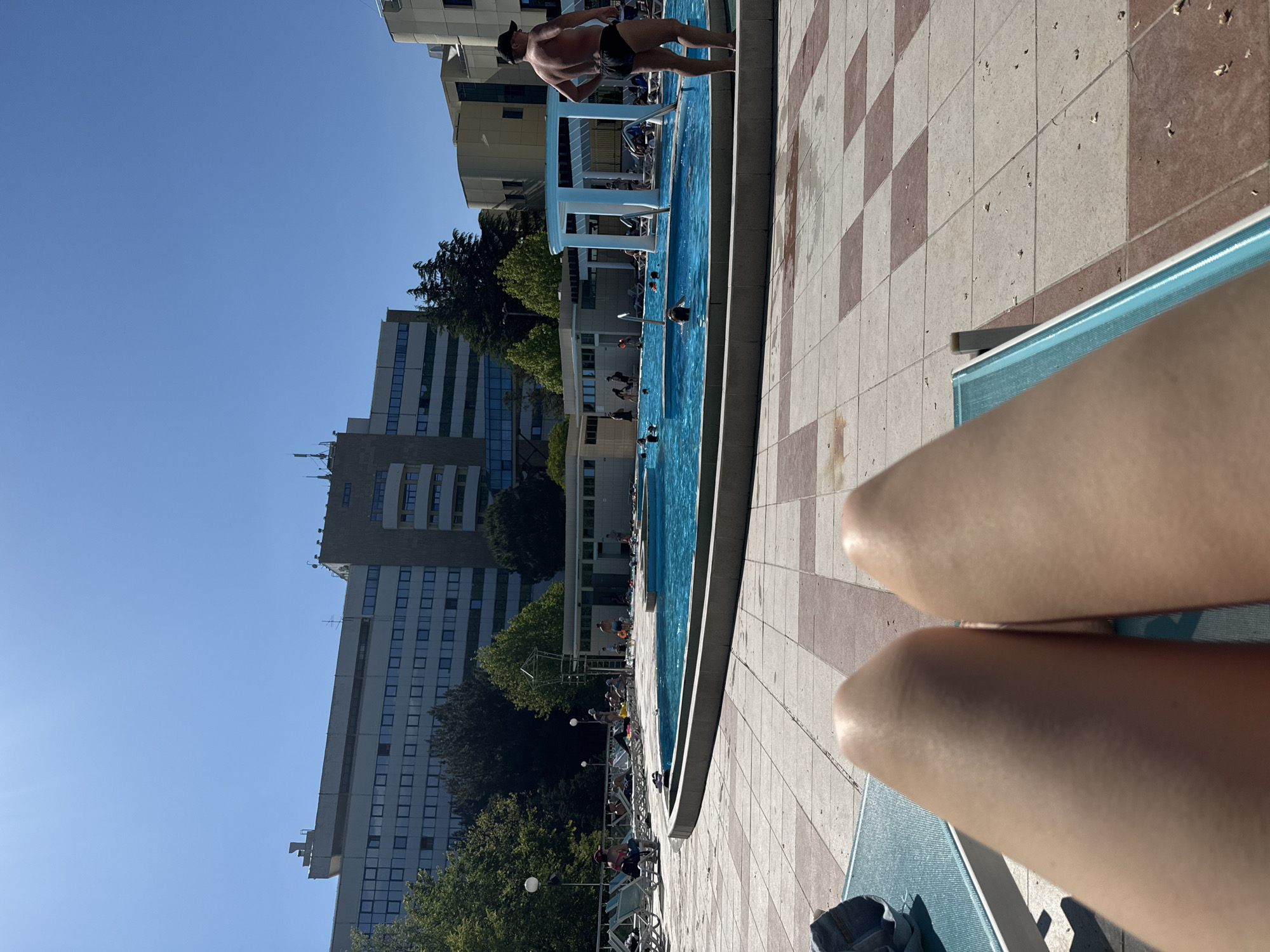 This image depicts an outdoor swimming pool area in Nebug, Russia, on a clear, sunny day. The perspective is from a person lying on a blue mesh lounge chair, with their light-skinned legs and knees prominently in the foreground. To the left of the legs, a partial blue denim item is visible on the lounger.
The ground immediately around the lounge chair is paved with large square tiles in a checkerboard pattern, alternating between light beige and reddish-brown. Small, light-colored debris is scattered across these tiles.
In the midground, a light blue, irregularly shaped swimming pool is the central feature. Several individuals, including adults and children, are present in the pool, either swimming or standing in the water. To the immediate right of the pool, an adult male with a light skin tone, wearing black swim briefs and a dark baseball cap, stands with his hands on his hips, facing away from the camera and towards the pool.
Around the pool, numerous other people are visible, some relaxing on lounge chairs, others walking or standing. A white, columnar pergola structure stands by the pool's edge, and other white, low-rise buildings (likely facilities) are present in the background of the pool area. A metal lifeguard-style chair or observation platform is also visible near the left side of the pool.
The background is dominated by a large, multi-story building, approximately 8-10 stories high, with a light-colored facade and numerous dark, rectangular windows arranged in horizontal bands. Architectural structures, including what appear to be vents or mechanical units, are visible on its roofline. To the right of this main building, another structure with a darker, textured facade and curved or rounded architectural elements is partially visible. Green deciduous trees with full foliage are situated around the pool area and in front of the background buildings. The sky is clear and bright blue, indicating strong sunlight, which casts distinct shadows.
