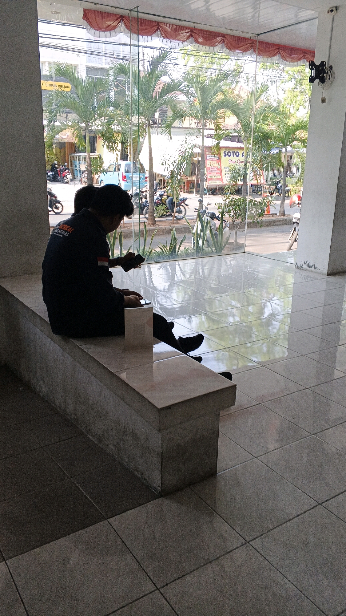 This image depicts the exterior of a building with a large glass window, likely located in Semarang, Indonesia, as suggested by the presence of palm trees and motorcycles commonly seen in Southeast Asian cities. Inside, two individuals are seated on a concrete bench. The person on the left is wearing a dark uniform with an emblem and is holding a smartphone, appearing to be engaged with it. Their legs are crossed, and they are wearing dark shoes. A white box is placed on the bench in front of them. The second individual, partially visible to the right, also appears to be looking at a smartphone. The floor inside the building is tiled and reflective, showing the reflection of the outside environment. Outside the glass, there are palm trees, a street with parked and moving motorcycles, and buildings with signs, including one that reads "SOTO AYAM". The overall atmosphere suggests a public space, possibly a waiting area or entrance to a business.