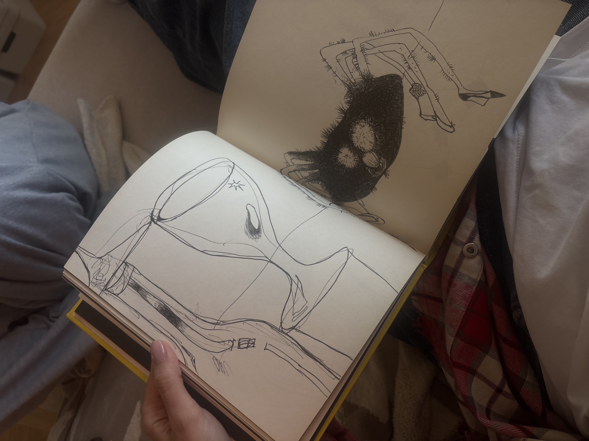 An open book displays two pages of ink drawings. The left page shows a stylized wine glass and bottle. The right page contains a drawing of a dark, fuzzy creature with legs wearing shoes. The book is held open by a hand with painted nails, on a lap of someone wearing jeans, a white shirt, and a red plaid jacket. The background consists of soft furnishings in neutral tones. The location is unknown and not necessarily in Krasnoyarsk, Russia.