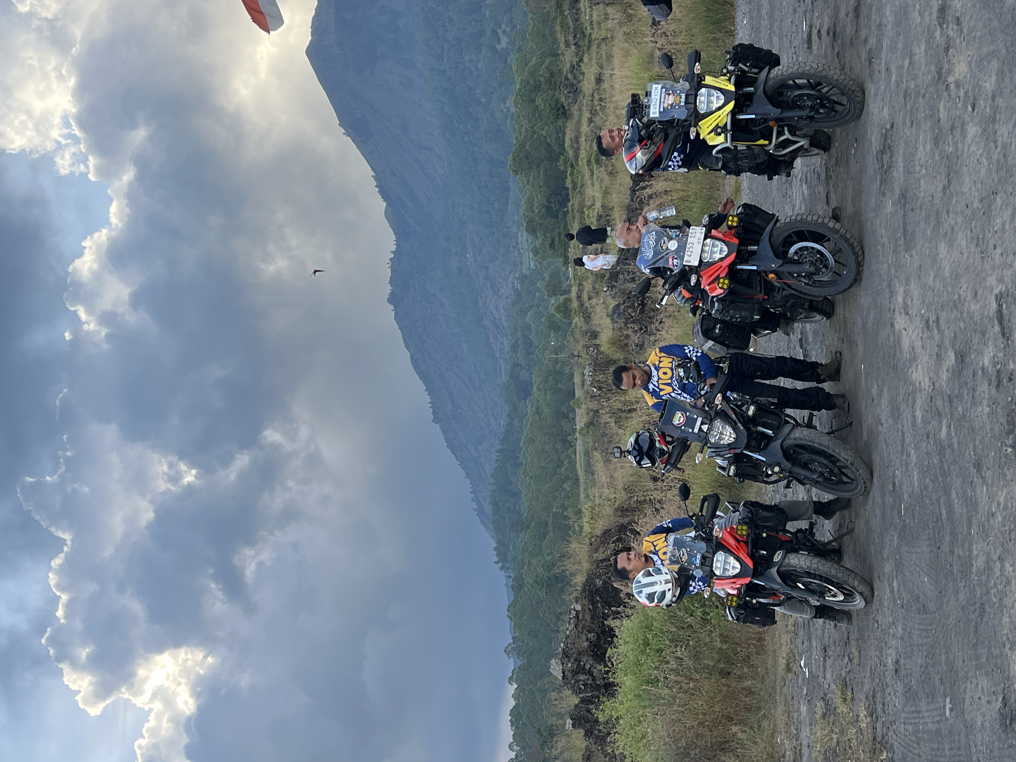 The image depicts four people and four motorcycles parked on a road with a mountain in the background under a cloudy sky. A paraglider is visible in the sky. The location is Kintamani, Indonesia. The motorcycles are adventure bikes, and the riders are wearing riding gear. They appear to be taking a break on a motorcycle tour.