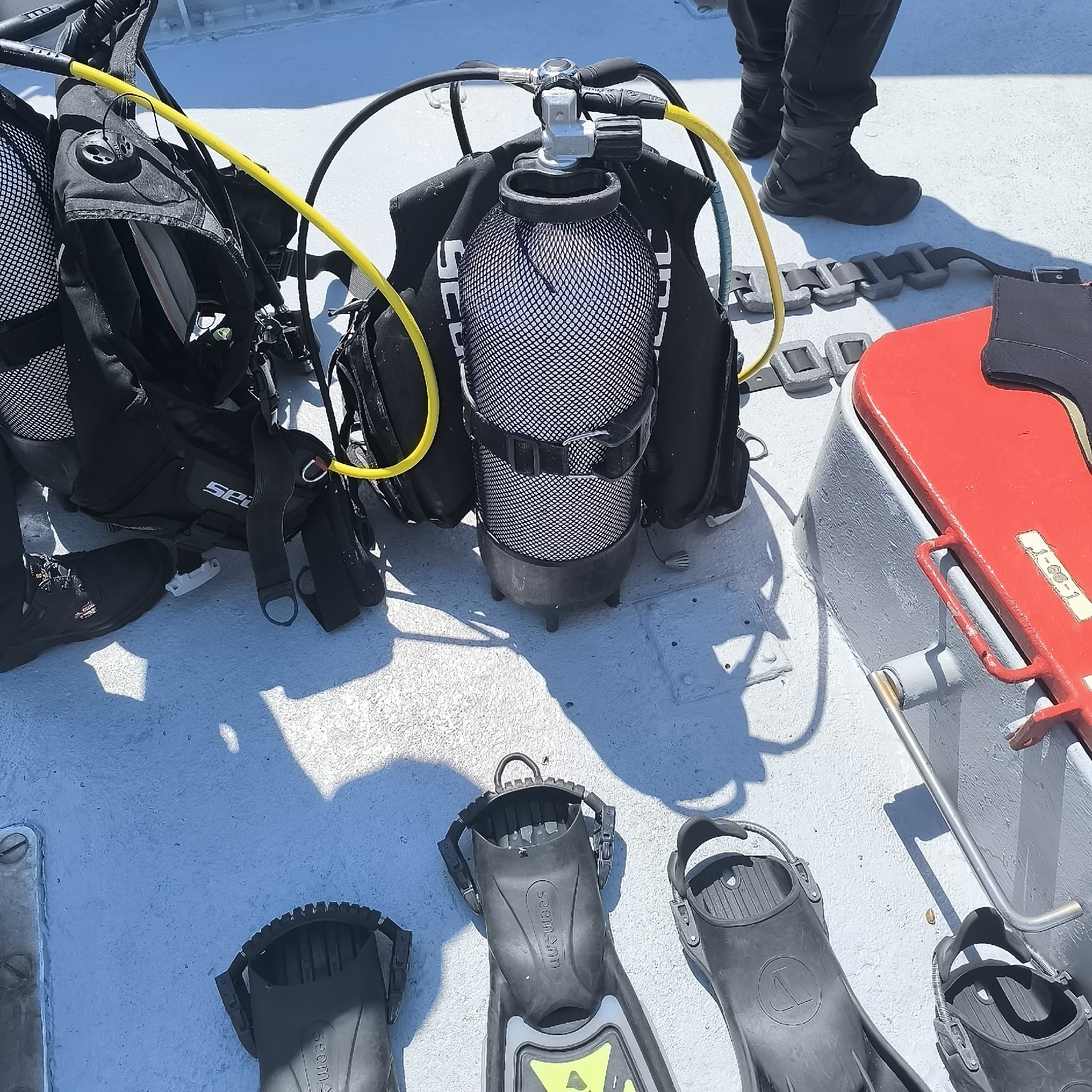The image shows a collection of scuba diving equipment laid out on a deck. The equipment includes at least two black buoyancy control devices (BCDs) with attached regulators and hoses, one of which has a white mesh cover over a scuba tank. There are also several black dive fins and a weight belt with rectangular lead weights. In the background, a person wearing black pants and tactical boots stands near the equipment. A portion of a red container is visible to the right of the image, with a metal handle. The deck appears to be painted a light gray color and is exposed to sunlight, casting shadows from the equipment and the person. The setting suggests an outdoor location, likely on a boat or pier, suitable for diving activities. There is no indication of the specific location within the city of Varna, Bulgaria.