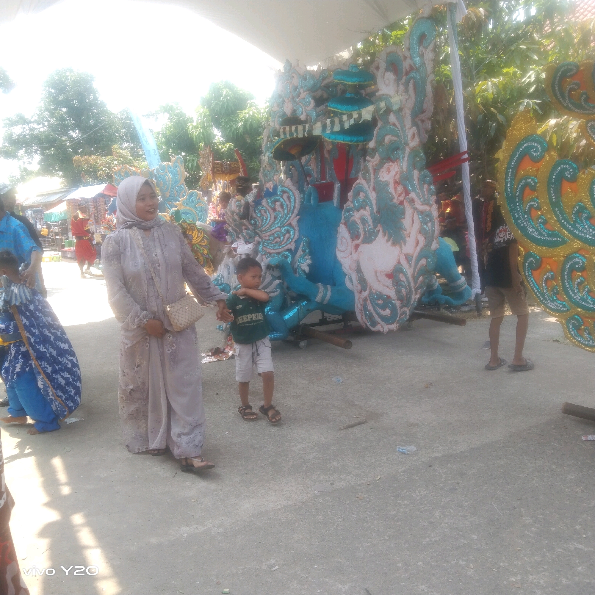 The image depicts an outdoor scene likely during a festival or parade, with brightly decorated floats. In the foreground, a woman wearing a light-colored hijab and a long, patterned dress stands with a small, tan purse slung across her body. To her left, a young boy in a green t-shirt and light shorts stands with his arms crossed. Behind them, a large, elaborate float with blue and white decorative elements is visible. Further to the left, another float with a yellow and teal design is partially visible. In the background, several other figures are present, including individuals in brightly colored clothing, suggesting a lively event. The ground is paved, and trees and buildings can be seen in the distance, indicating an urban or semi-urban setting. The sky is bright, suggesting daylight, with some glare from the sun.