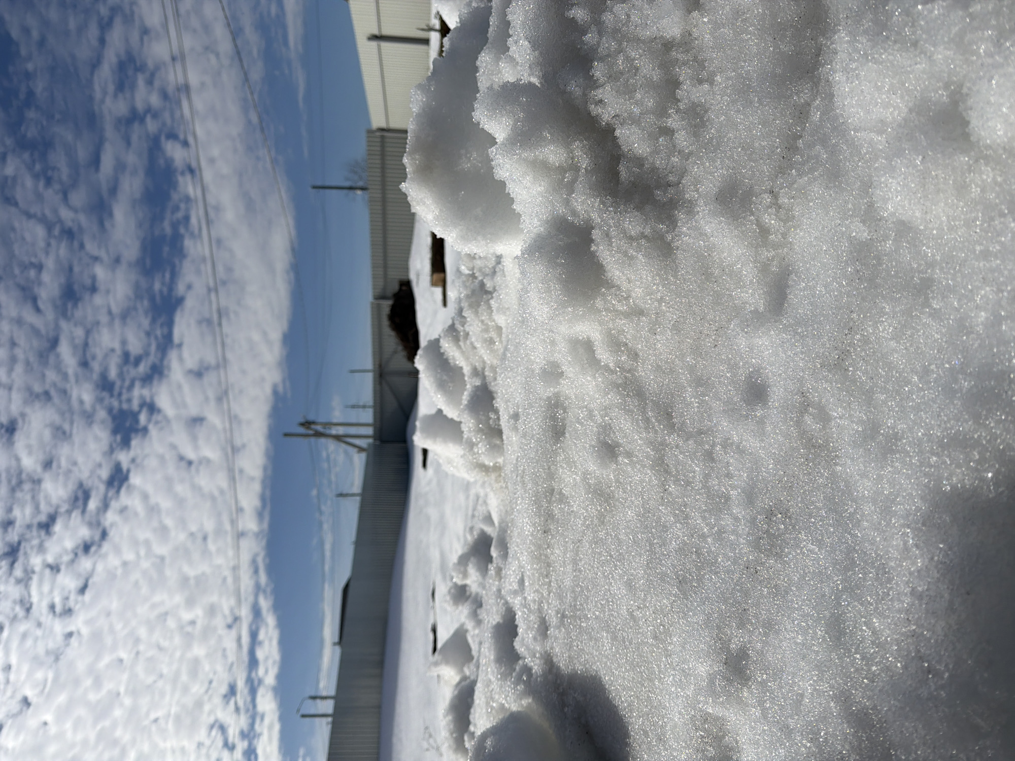 This image depicts a snow-covered outdoor area in Chaikovsky, Russia, under a bright, partly cloudy sky. The foreground is dominated by uneven mounds of crystalline, glistening snow, suggesting recent snowfall or partial melting and refreezing. The ground beyond the immediate foreground is also covered in snow. In the midground, a dark pile of what appears to be soil or debris is visible, along with some wooden planks or beams resting on the snow. The background features a continuous light-colored metal fence, likely constructed from corrugated or sheet metal panels, enclosing the space. Above the fence line, multiple utility poles, one equipped with a light fixture, and associated power lines are observed stretching horizontally across the upper portion of the frame. Bare tree branches are faintly visible near one of the utility poles. The sky is blue with scattered white altocumulus clouds. No individuals or active subjects are present in the scene. The lighting indicates direct sunlight, casting subtle shadows and highlighting the reflective qualities of the snow.