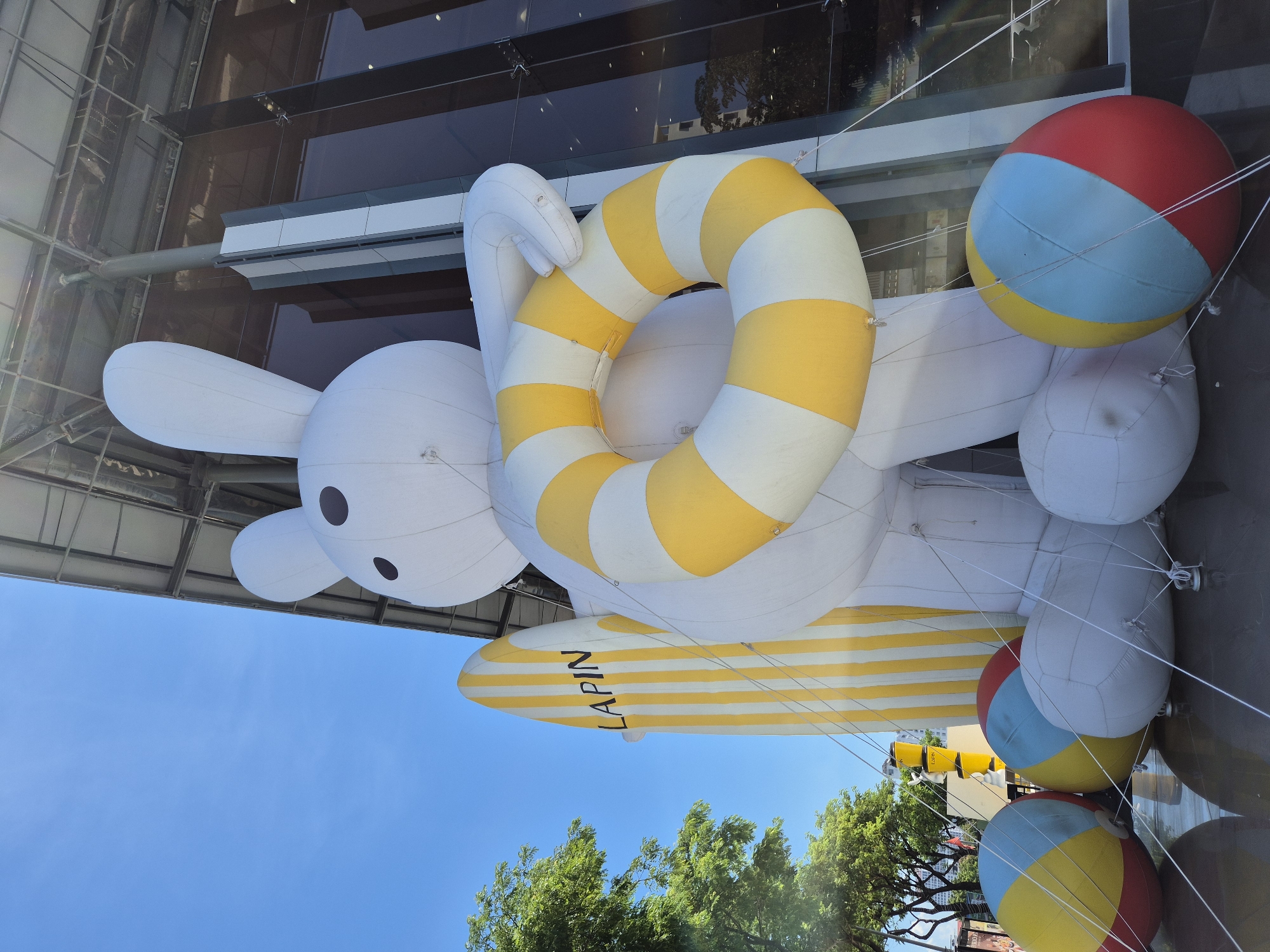 A giant inflatable white bunny stands outdoors, holding a yellow and white striped inner tube and a yellow and white striped surfboard that reads "LAPIN". The bunny is positioned in front of a modern building with large glass windows. Several large, colorful beach balls are placed at the base of the inflatable. The scene suggests a festive or promotional event. The presence of trees and clear blue sky indicates an outdoor location, likely in a city. The image was taken in Da Nang, Vietnam.