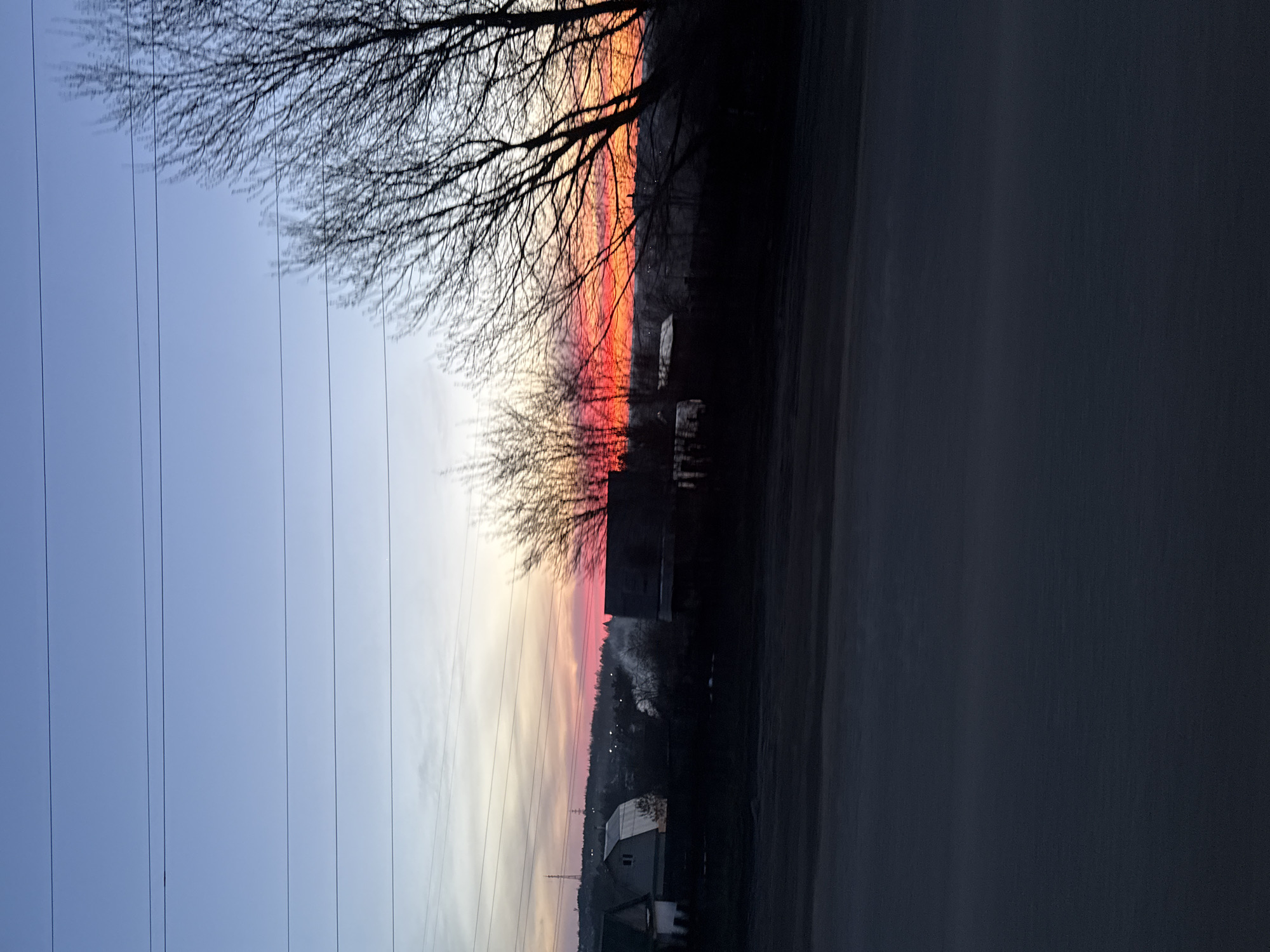 The image shows a sunset scene in what appears to be the outskirts of a town, possibly Chaikovsky, Russia. The sky exhibits a color gradient from a dark blue at the top, transitioning to orange and red near the horizon. Silhouetted bare trees and power lines are visible in the upper portion of the frame. In the lower section, there's a blurred dark surface, likely a road or pathway. Further into the distance, one can see a row of houses and structures, with smoke rising, suggesting the presence of a chimney. The lighting and colors indicate either sunrise or sunset.