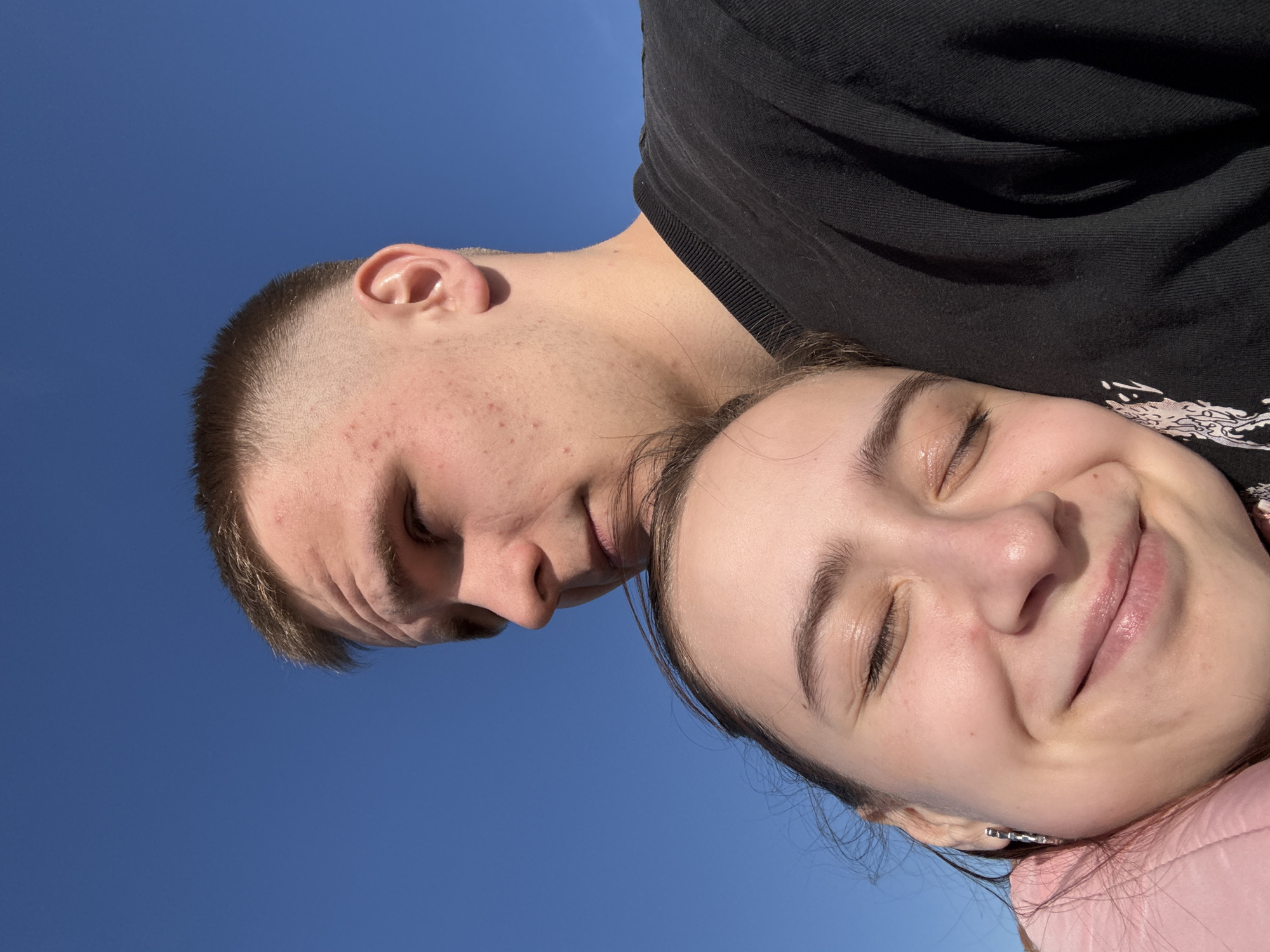 The image displays two young individuals, a male and a female, positioned closely together outdoors against a striking, clear blue sky. The photograph is oriented 90 degrees clockwise, making the subjects appear to be lying down or captured from a tilted perspective.
The female subject, closer to the viewer and on the right side of the frame, has her eyes gently closed and a clear, serene smile on her face, conveying contentment or happiness. She is wearing a pink garment, possibly a jacket or top, and a subtle earring is visible. Her head seems to be resting on the male subject's shoulder or chest.
The male subject, partially obscured behind the female and on the left, is wearing a dark, possibly black, t-shirt or sweatshirt. He has very short hair and his face, which shows some visible acne, is angled downwards with his eyes also mostly closed or looking away.
The strong, direct sunlight on their faces and the absence of clouds suggest a bright, sunny day, likely midday or afternoon. The background is an unblemished blue sky, indicating an open outdoor setting without specific landscape features. No visible text is present in the image. The scene is set in Votkinsk, Russia.
