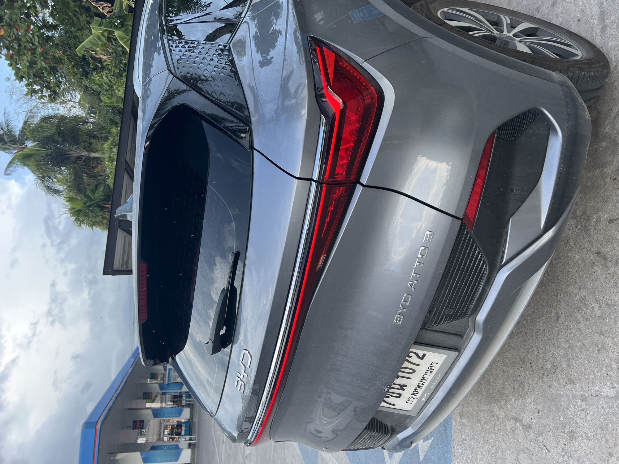 The image shows the rear of a gray BYD ATTO 3 car, with a Thai license plate visible. The car is parked at a gas station, and in the background, tropical vegetation is visible. The location is likely in or near Baan Bo Phut, Thailand, as indicated on the license plate frame. No people are visible in the shot.