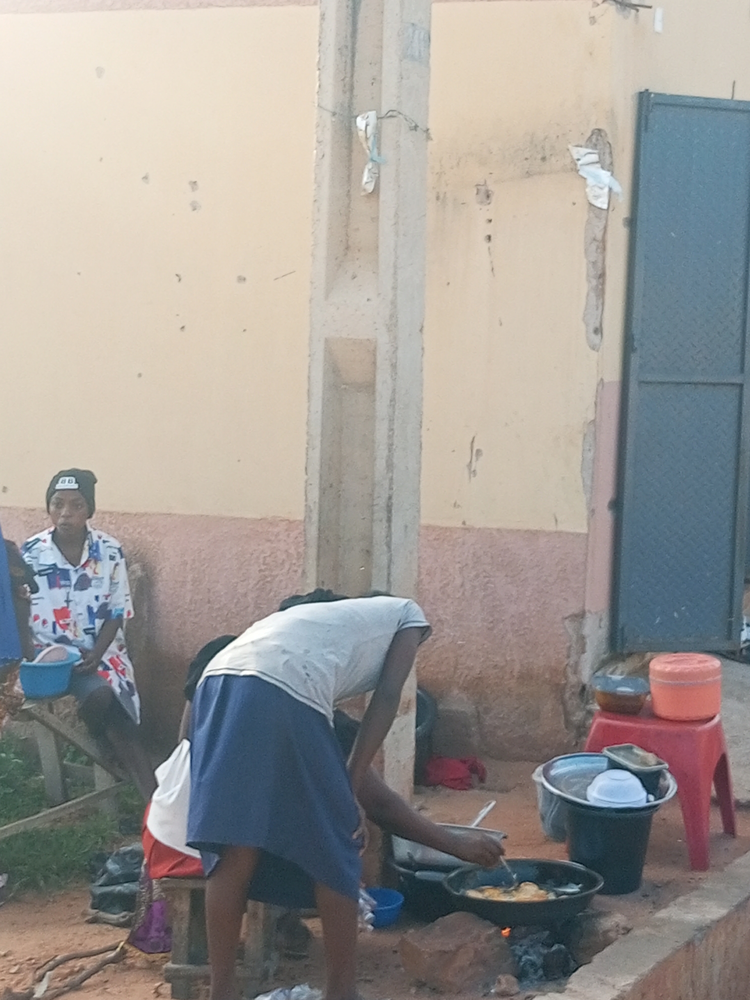 In Gombe, Nigeria, a scene unfolds with an outdoor cooking setup. A person, wearing a white t-shirt and a blue skirt, is bent over a pot placed on a makeshift stove. Flames are visible beneath the pot, indicating cooking is in progress. The person is holding a utensil, seemingly stirring or tending to the food. To the left, another individual sits on a wooden structure, wearing a patterned shirt and a black beanie. They are holding a blue bowl. In the background, a textured wall, painted in light yellow and pink, runs along the side of a building. A blue metal door is visible to the right. Several containers and bowls are arranged around the cooking area, including an orange container on a red stool. The ground is covered with dirt and debris.