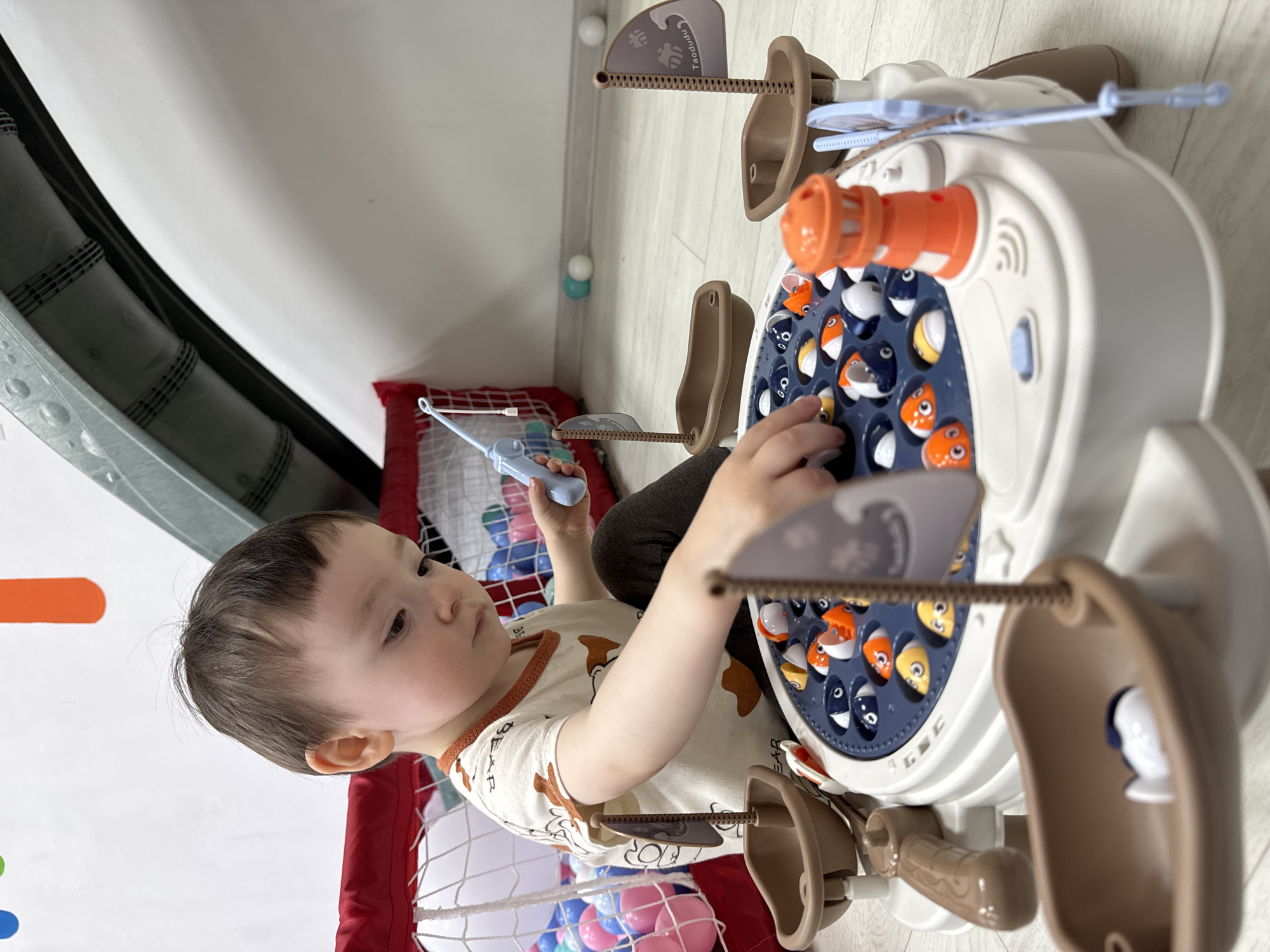 A toddler is playing with a toy fishing game inside a playpen filled with colorful plastic balls. The fishing game features rotating fish in a circular pattern. The child is holding a toy fishing rod. The scene takes place indoors on a light-colored floor next to a white wall.