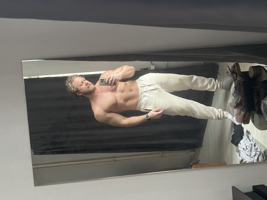 The image is a mirror selfie taken indoors, depicting a shirtless, muscular male with curly blond hair and a beard, facing a large rectangular mirror. He wears light beige sweatpants; his left foot is covered by a grey sock, while his right foot is bare. A dark-colored smartwatch is present on his left wrist. He holds a silver-colored smartphone in his right hand, angled to capture his reflection, and looks downward with a slight curve to his mouth. A nipple piercing is visible on his left nipple. The background reflected in the mirror consists of a dark grey or black curtain directly behind the man and a light-colored wall to his left. In the foreground of the actual room, a pile of crumpled clothing, including a dark shirt, lies on the floor. Adjacent to the clothing is a small, dark wooden table or stand holding various items, including cylindrical containers and a white object partially inscribed with "SPRIT." The indoor setting does not provide specific external architectural details or landmarks to identify a precise location within the city of Saint Petersburg, Russia.