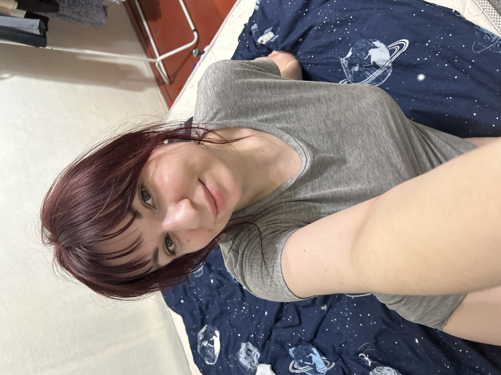 A woman is lying on a bed with a blue comforter featuring a planet and star pattern. She is wearing a gray t-shirt and her hair is burgundy. The background includes a wall and a white shelf. The location, Фоки, Russia, cannot be determined from the image.