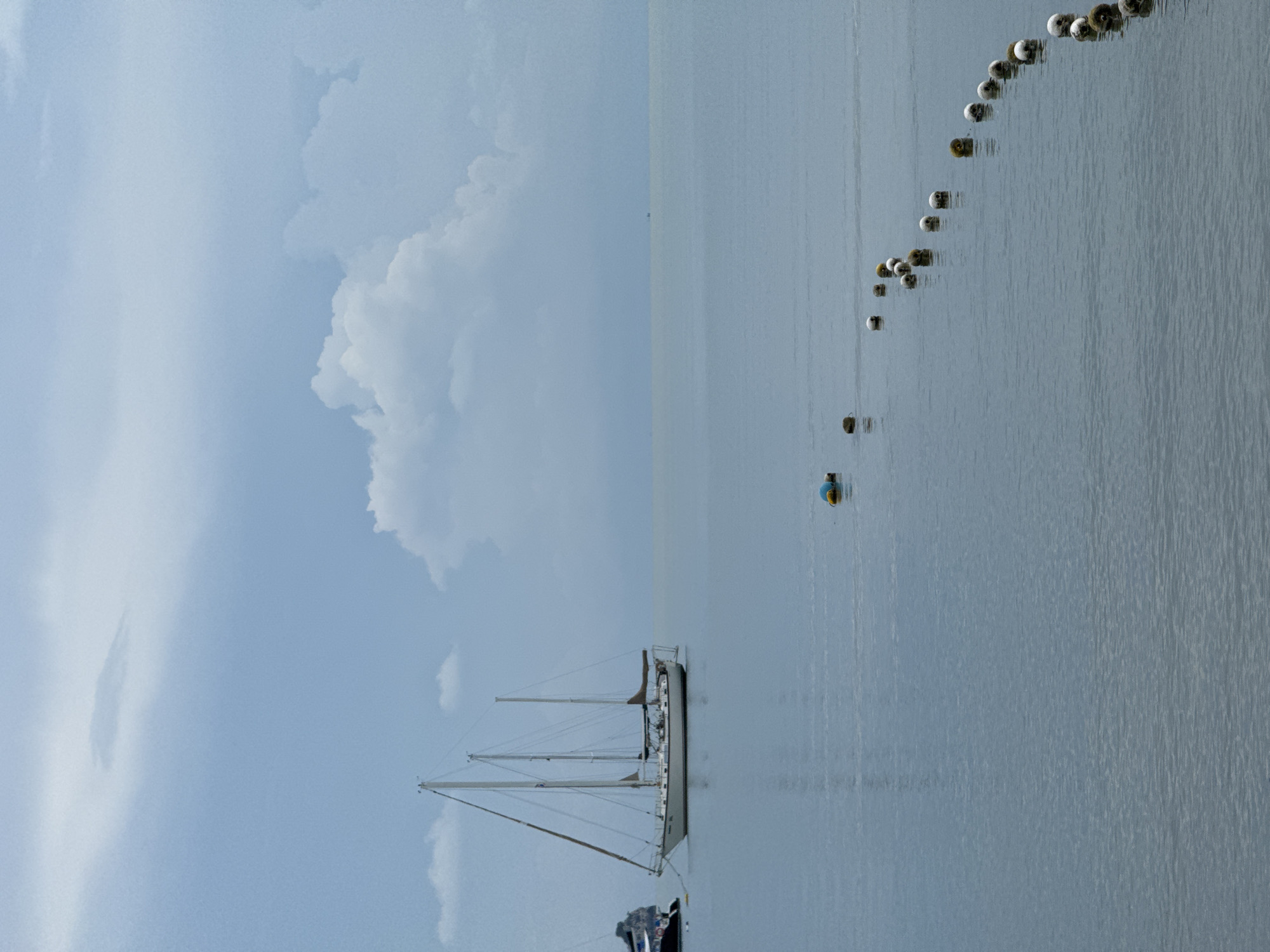 This image depicts a serene seascape with a prominent sailboat anchored in calm, light blue water. The sailboat, a white hull with three masts and rigging, is positioned to the left of the frame. A portion of another dark-hulled boat is visible to the far left. A line of white, yellow, and green buoys stretches diagonally across the lower right portion of the image, marking a boundary or path in the water. The sky is overcast with soft, grey clouds, and a large, puffy white cloud is visible towards the center-right. The horizon line is faint, suggesting a hazy or distant shoreline. The overall atmosphere is tranquil and quiet. The location is described as Unknown, Unknown.