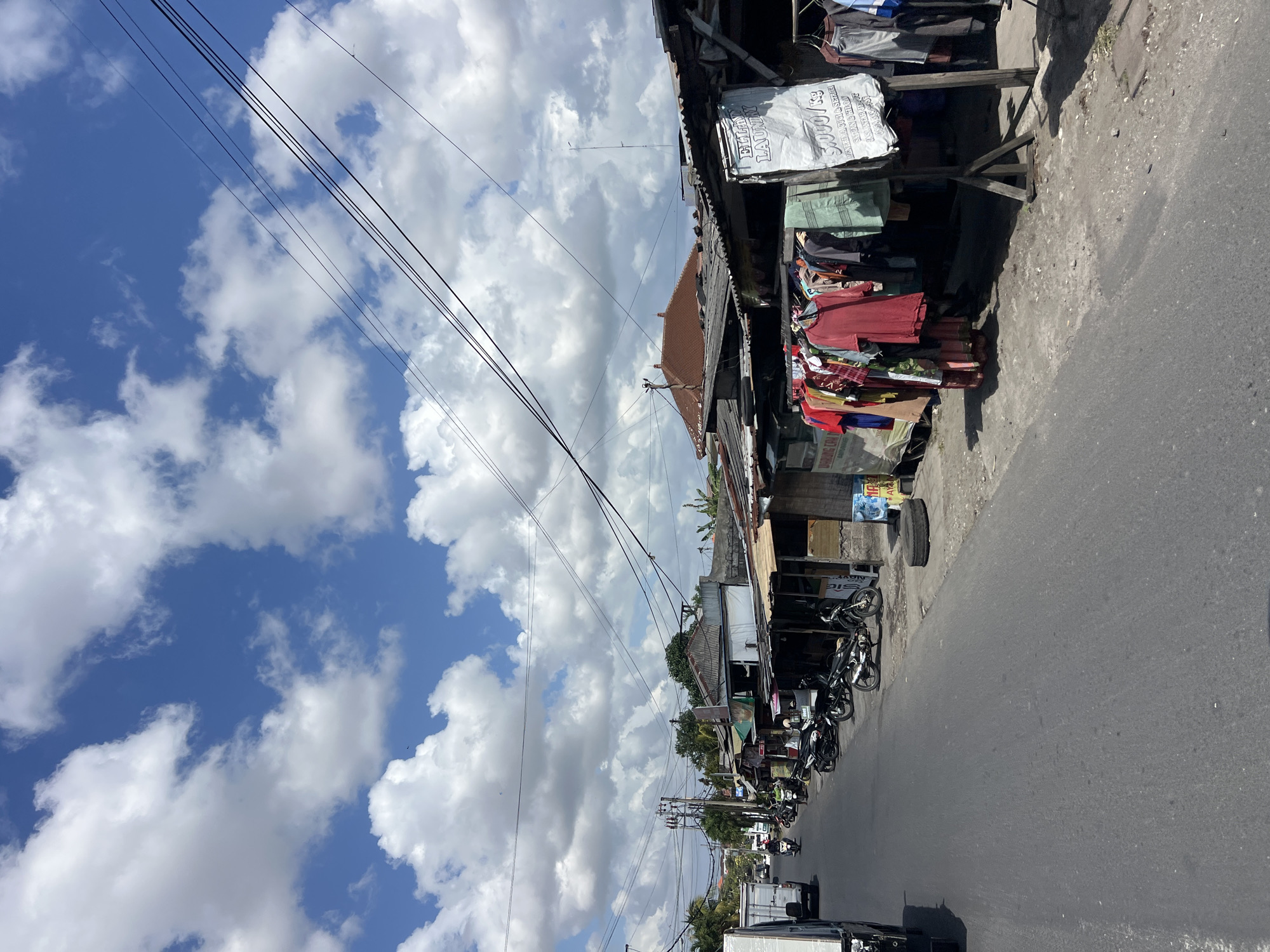 The image depicts a street scene in Denpasar, Indonesia, with a focus on a row of small shops and stalls lining the road. The sky is a bright blue with scattered white cumulus clouds. Overhead, a network of black electrical wires crisscrosses the frame.
On the right side of the street, several open-fronted stalls are visible, displaying a variety of clothing items hung on racks. One stall has a large white sign with text, possibly advertising laundry services, with "5,000/kg" prominently displayed. Next to this stall, a black tire rests on the ground. The stalls themselves appear to be constructed with corrugated metal roofs and wooden frames.
Along the left side of the asphalt road, a line of motorcycles is parked. Further down the road, a white truck is in motion, and other vehicles and motorcycles can be seen in the distance. The shops and stalls are simple structures, likely operating as informal businesses. The environment appears to be a typical urban or semi-urban street in a tropical climate, characterized by the presence of motorbikes, roadside commerce, and overhead utility lines.
