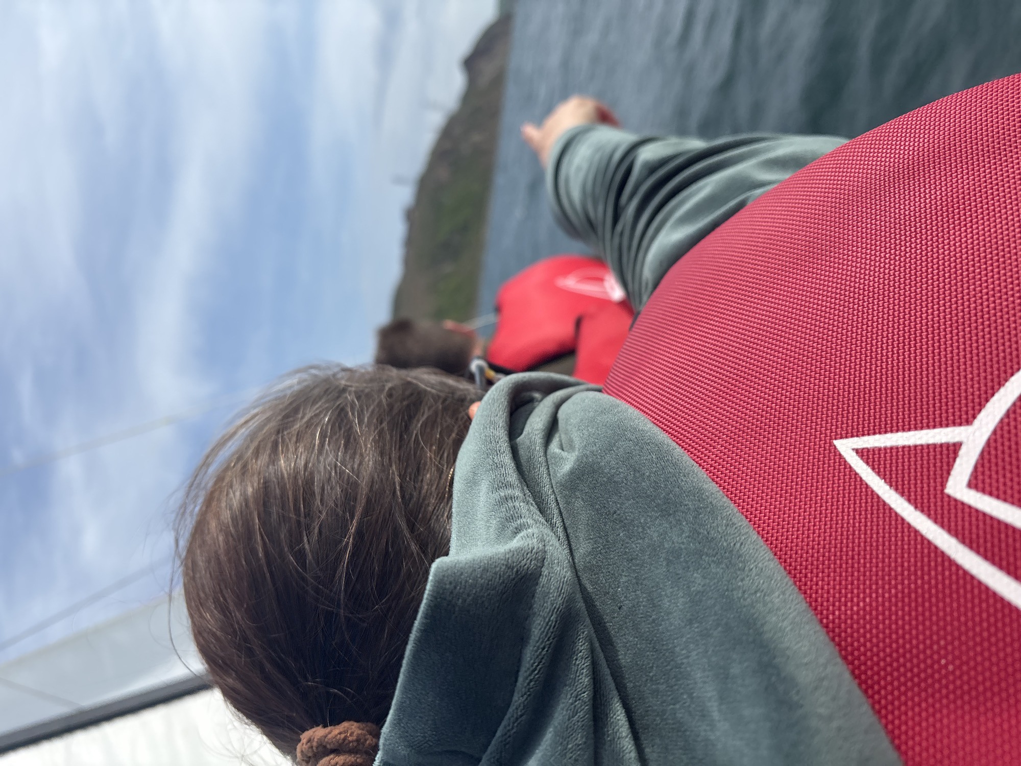 A slightly angled view captures a person with brown hair and a dark green jacket pointing towards an island. The person is wearing a red life vest with a white logo. In the background is a body of water with a glimpse of the sky and clouds. The specific location of Vladivostok, Russia is not clearly discernible from the details available in the image.