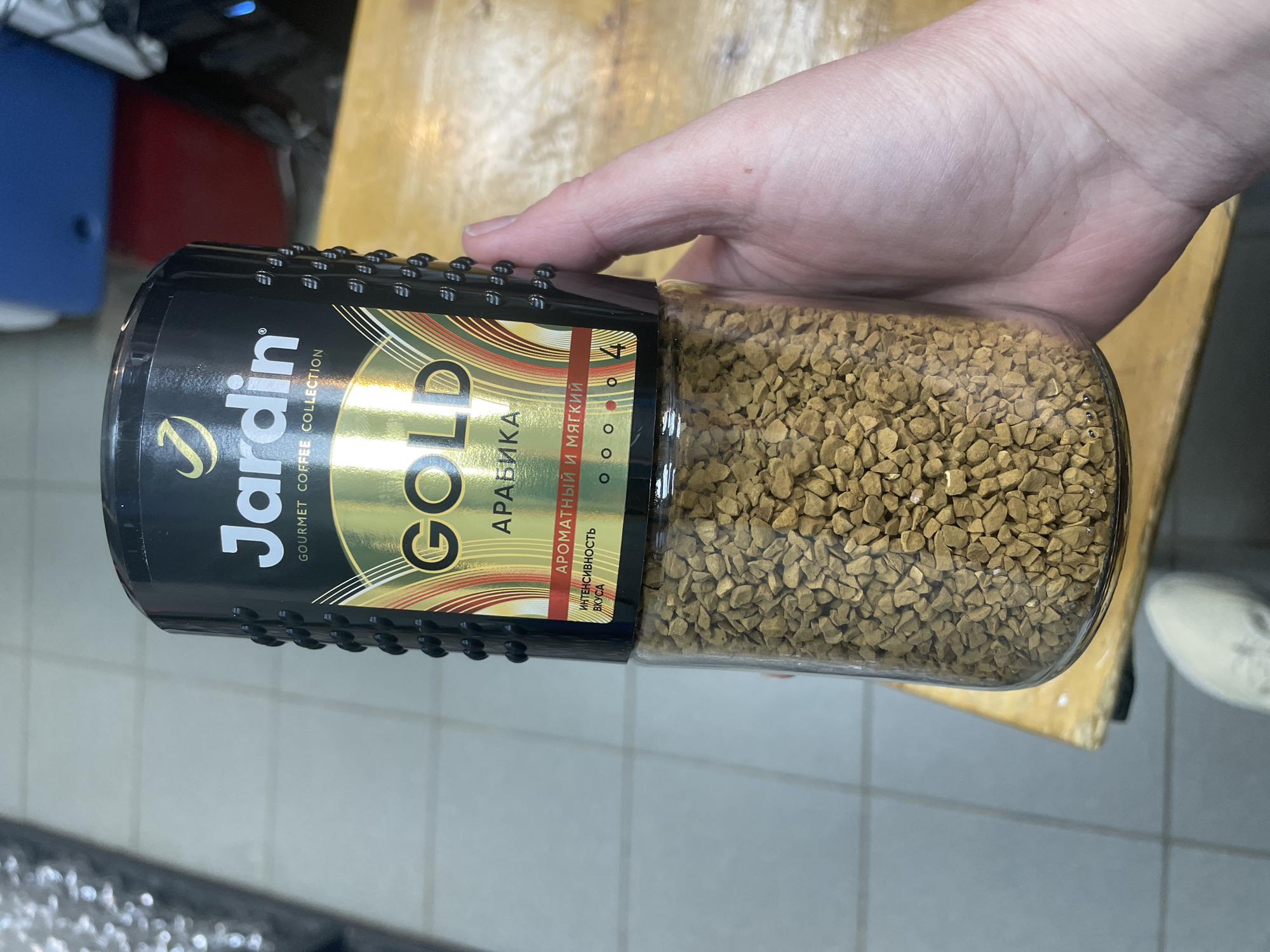 A hand is holding a glass jar filled with instant coffee granules. The jar has a black plastic lid and a label with gold and black accents. The label prominently displays the brand name "Jardin" and the word "GOLD" in large, stylized letters. Below "GOLD," the word "АРАБИКА" is visible, indicating the coffee type. Further text on a red rectangular section reads "АРОМАТНЫЙ И МЯГКИЙ" which translates to "AROMATIC AND SOFT" and below that "ИНТЕНСИВНОСТЬ ВКУСА" meaning "INTENSITY OF TASTE." The setting appears to be indoors, with a wooden surface beneath the jar and a tiled floor visible in the background. The lighting suggests it is daytime. The image's orientation is rotated 90 degrees clockwise.