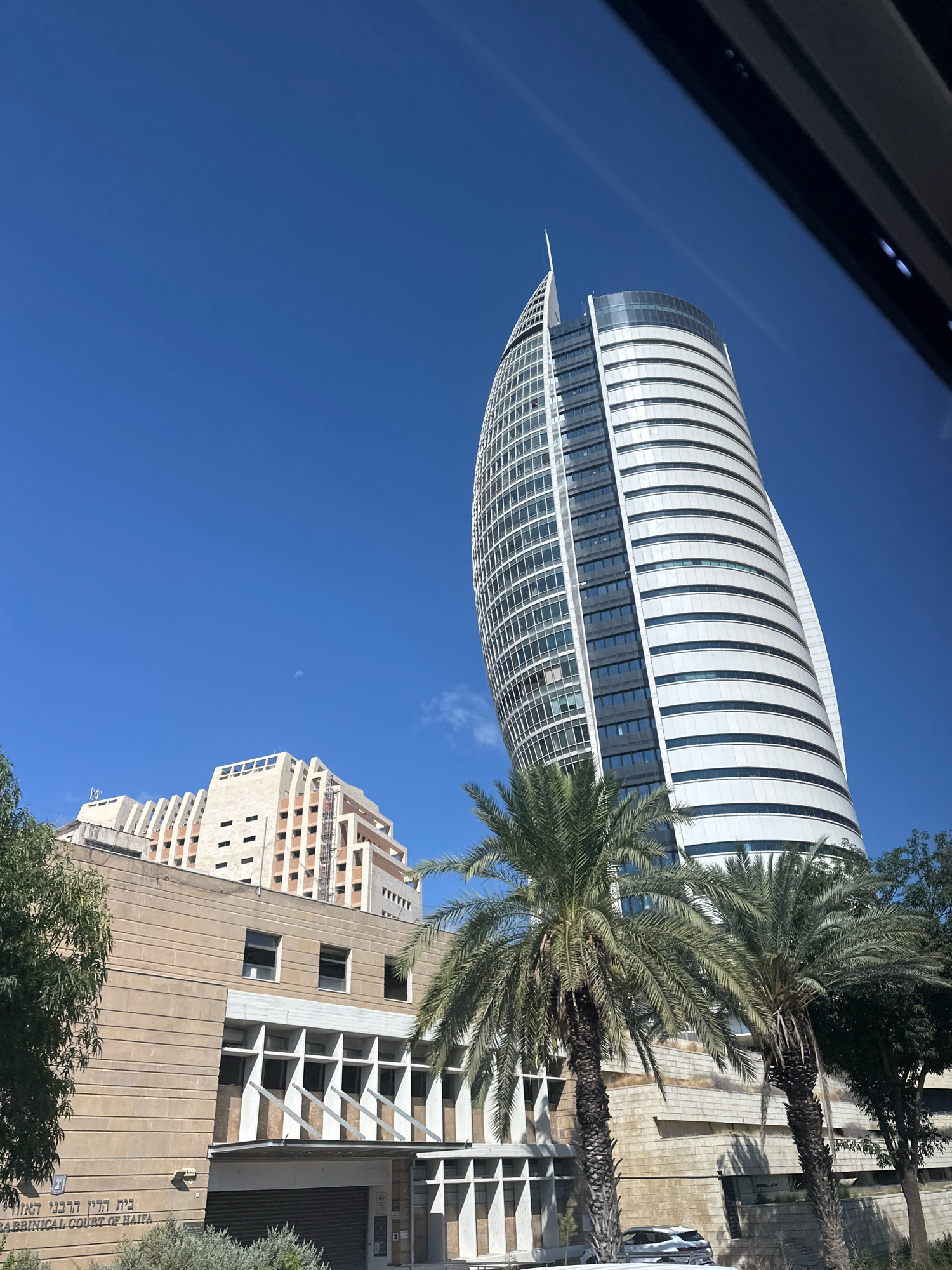 The image captures a low-angle view of modern architecture against a clear blue sky in Haifa, Israel. Dominating the right side of the frame is a tall, spiraling skyscraper with a white, ribbed facade and extensive glass windows. In the background, to the left of the skyscraper, stands a tan, stepped building with numerous windows and a patterned facade. In the foreground, a lower, tan stone building with a modernist design features a prominent entrance area with supporting pillars and a dark, closed roller shutter. The signage on this foreground building reads "בית הדין הרבני הראשי" and "RABBINICAL COURT OF HAIFA" in Hebrew and English respectively, identifying it as the Rabbinical Court of Haifa. Several palm trees are strategically placed throughout the scene, with their fronds framing parts of the buildings and adding a touch of nature to the urban landscape. A sliver of what appears to be a vehicle is visible at the bottom of the image. The overall impression is one of a bright, sunny day in a city with contemporary and established architectural styles.