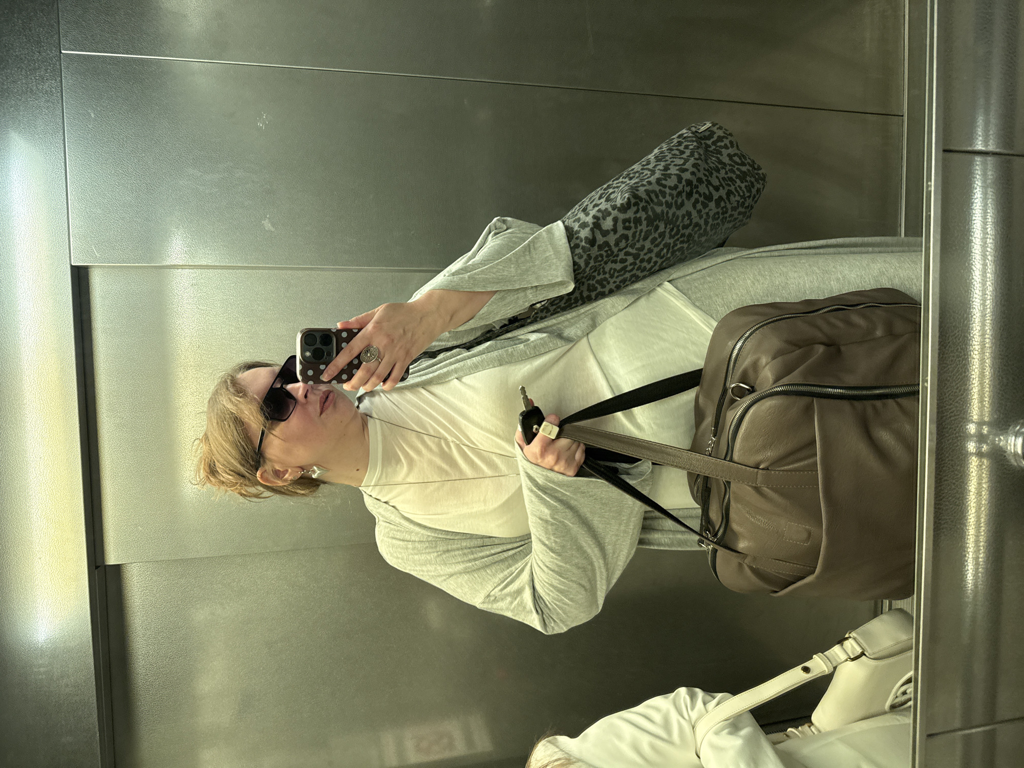 A woman with blonde hair, wearing sunglasses, a white t-shirt, and a grey cardigan, is taking a selfie in a metal-walled elevator. She is holding a phone in her left hand and a key in her right hand. She has a large brown leather bag hanging from her shoulder, as well as a leopard print pillow or bag, and a small white bag visible in the mirror. The environment appears to be a modern building. The location in Izhevsk, Russia, is not evident from the image.