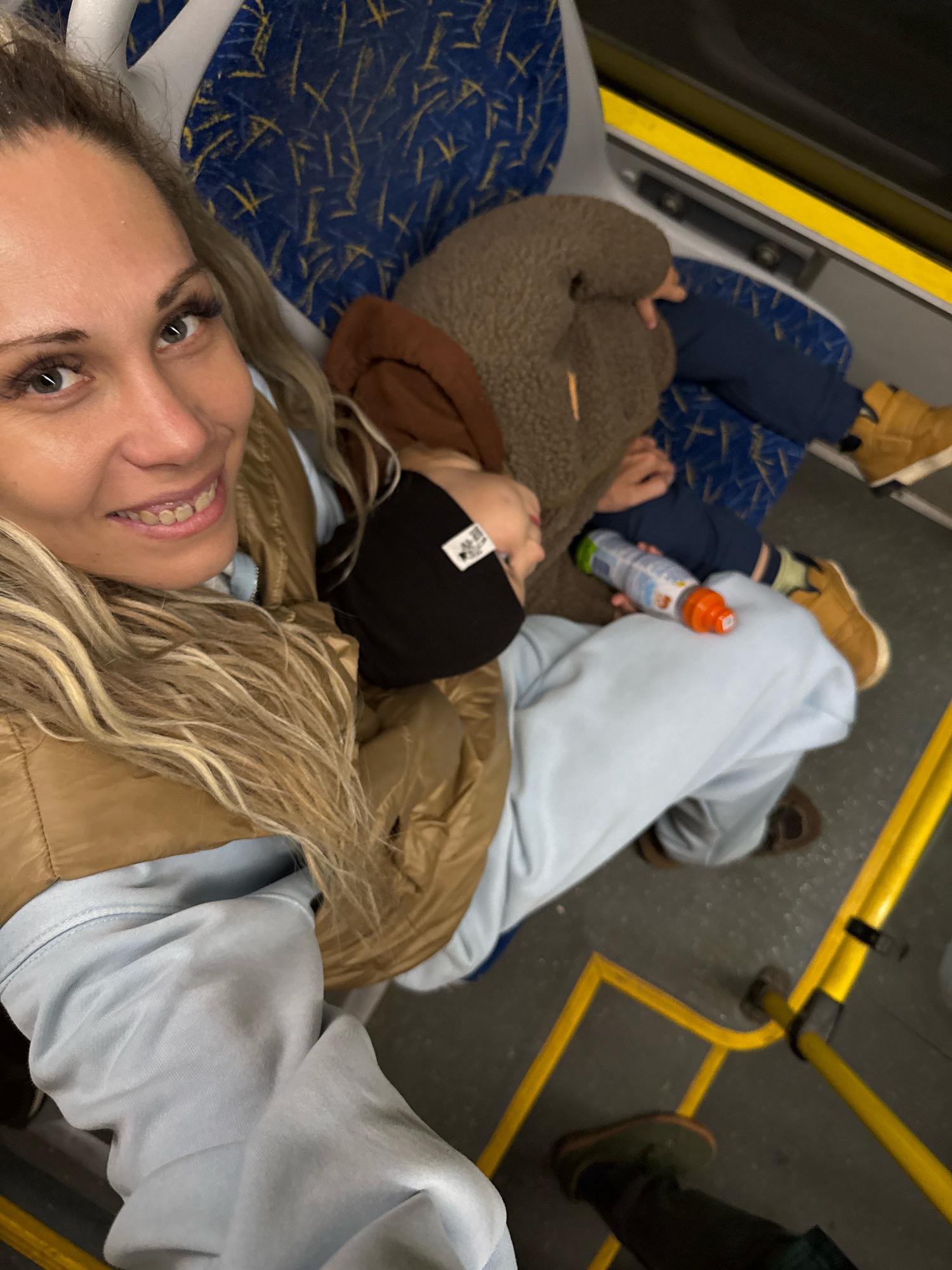 A woman with blonde hair and false eyelashes is taking a selfie on a bus, smiling and looking at the camera. She is wearing a light blue hoodie and a tan puffer vest. A child is asleep on her lap, wearing a brown hooded garment and a black cap. The child is holding a bottle with an orange cap and a label. They are seated on a bus with blue and yellow patterned seats. The bus has yellow handrails and the floor is dark gray. The location is identified as Rusanovo, Russia.