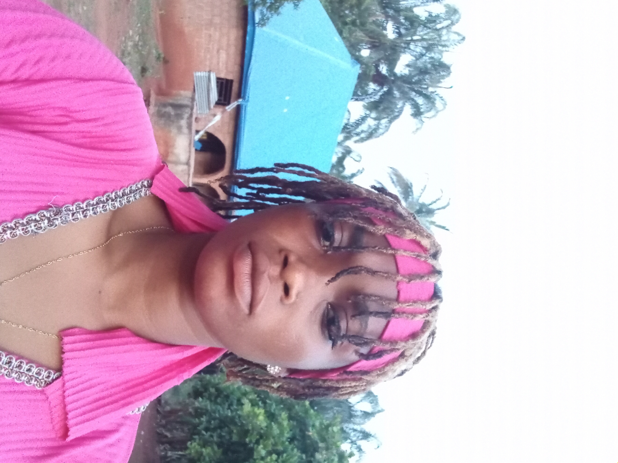 The image is a portrait of a woman, oriented vertically but rotated 90 degrees clockwise. A dark-skinned woman is prominently featured in the foreground, looking directly at the camera with a neutral, calm expression. She has full lips and her eyes are accentuated with dark eyeliner or mascara. Her hair is styled in dreadlocks, some of which appear to have pink highlights or are covered by a matching pink headwrap. She is wearing a bright, pleated pink top or dress with a silver-colored, decorative trim along the neckline. A thin, gold-colored chain necklace is visible around her neck.
The background depicts an outdoor setting during daytime, under what appears to be an overcast sky. Behind the woman, a building with a light blue roof and terracotta-colored walls is visible, along with some corrugated metal or scaffolding nearby. To her left and right, lush green trees and palm fronds are present, indicating a tropical or subtropical environment. The overall scene suggests a residential or semi-rural area in Ogbe, Nigeria, as provided by the context. No visible text is present in the image.