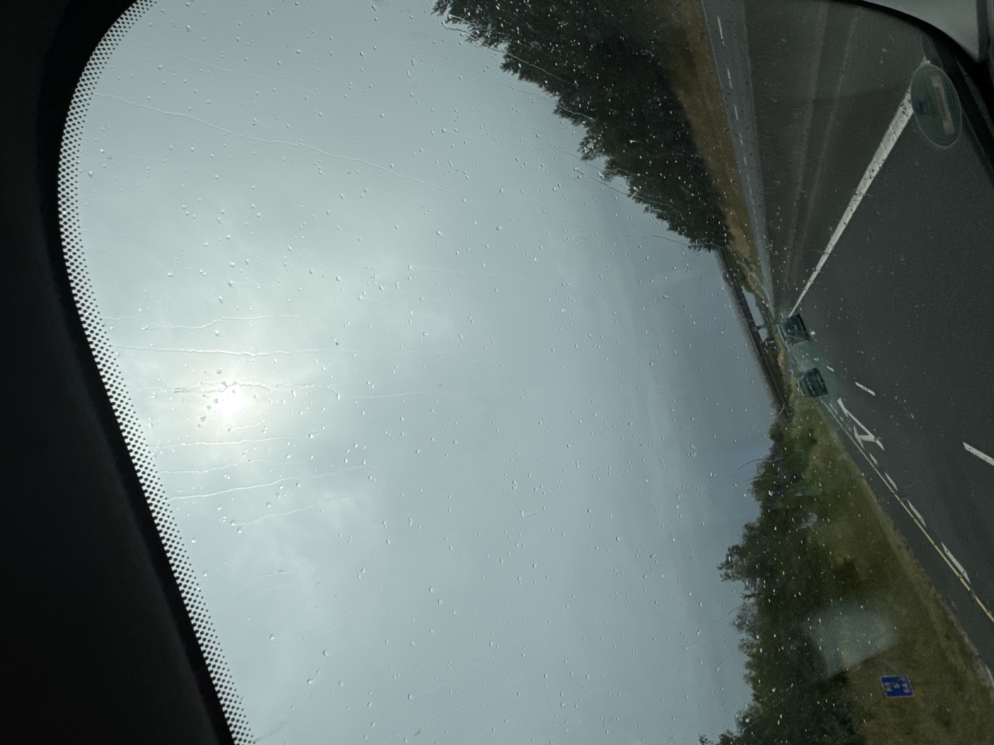 The image, captured from inside a vehicle, shows a view through a wet windshield on a highway. Raindrops are visible on the glass, distorting the scene. The sky is overcast with the sun faintly visible through the clouds. A dark line of trees borders the right side of the highway, and a concrete overpass can be seen in the distance. Two cars are traveling ahead on the road. The scene appears to be a rural highway, possibly in Ireland, given the mention of Lackabeg. There are no people clearly visible, only the interior of a car and the road ahead.