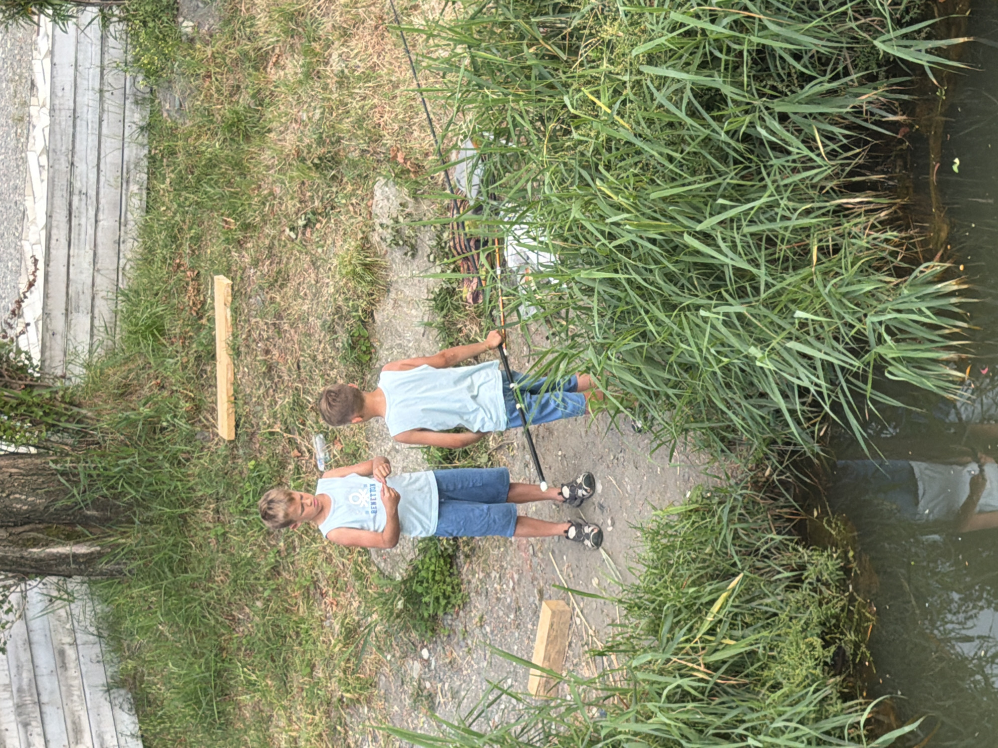 Two young boys are fishing by the water. The boy in the foreground is wearing a light blue tank top with the text "BENETTON" visible on it, and blue shorts. He is wearing sandals. The boy behind him is wearing a similar light blue tank top and blue shorts. Both boys are facing away from the camera, with their attention focused on the water. They are standing on a dirt path beside tall green reeds. The water appears dark and calm, reflecting the surrounding greenery and the sky. A fishing rod is held by the boy in the background, suggesting they are actively fishing. Behind the boys, a tree with rough bark is visible, and to their left, a wooden fence or siding runs parallel to a path made of concrete slabs. The scene appears to be outdoors on a sunny day, with the sunlight casting shadows. The overall mood seems relaxed and focused, typical of a fishing outing.