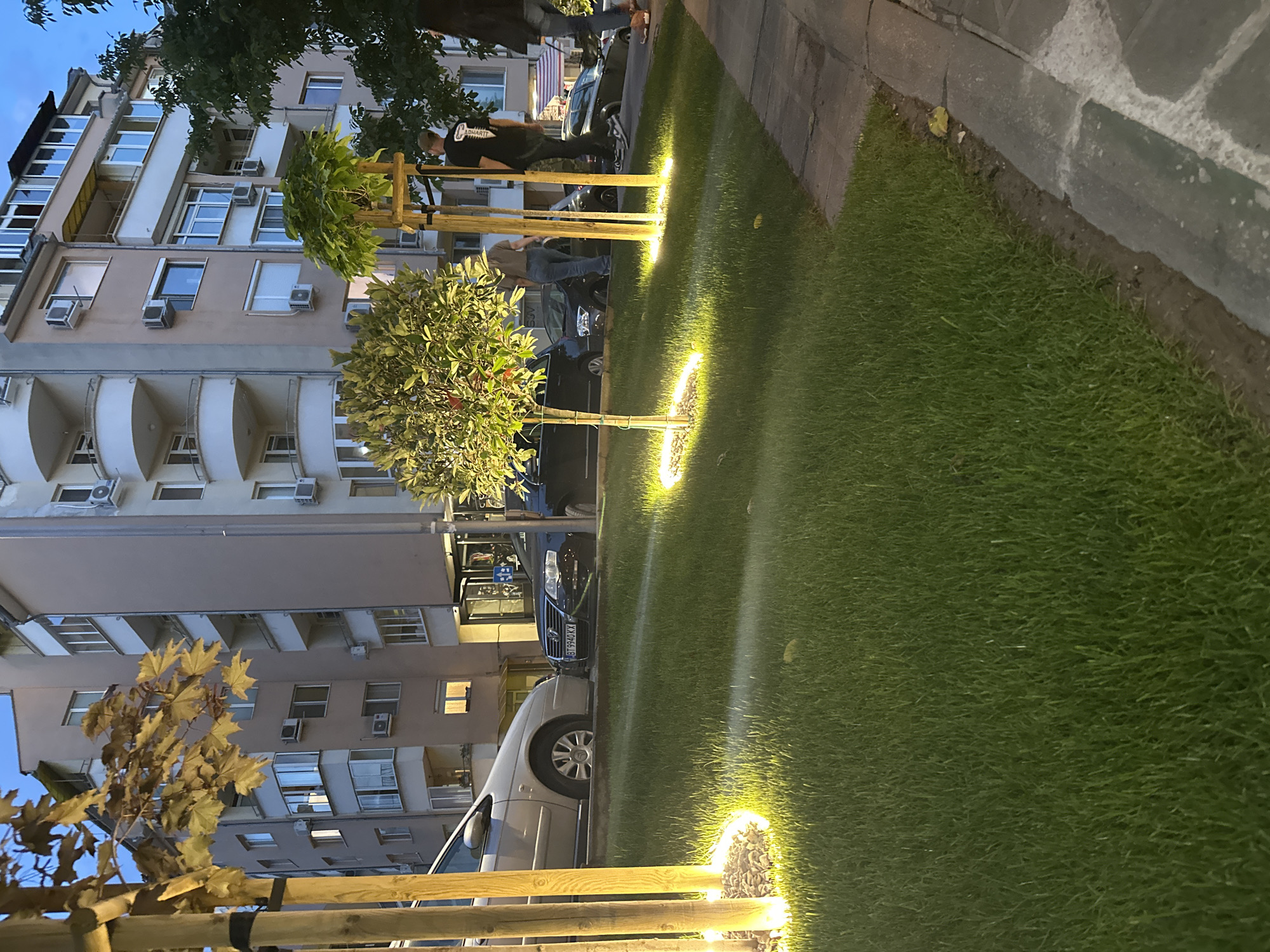 Here is a description of the image:
The image depicts a city street scene at dusk in what appears to be Veliko Tarnovo, Bulgaria. Tall buildings, possibly apartments, line one side of the street. Several parked cars are visible. The foreground features a landscaped area with lush green grass and small trees planted in regular intervals, each illuminated by a circular ground light. Wooden posts or beams are partially visible at the edge of the frame, possibly a part of a fence or other structure. People are walking near the trees. The overall atmosphere is calm and illuminated.