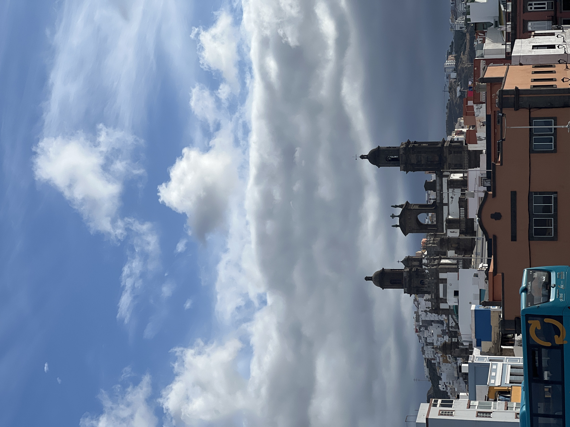 The image depicts the cityscape of Las Palmas De Gran Canaria, Spain. The sky is partly cloudy, with a clear blue section contrasting with a large, heavy cloud formation. In the foreground, the towers and facades of Catedral de Santa Ana are prominent. A partially visible blue bus is also noticeable on the right-hand side of the frame. Buildings and residential areas stretch into the background, gradually fading into the horizon.