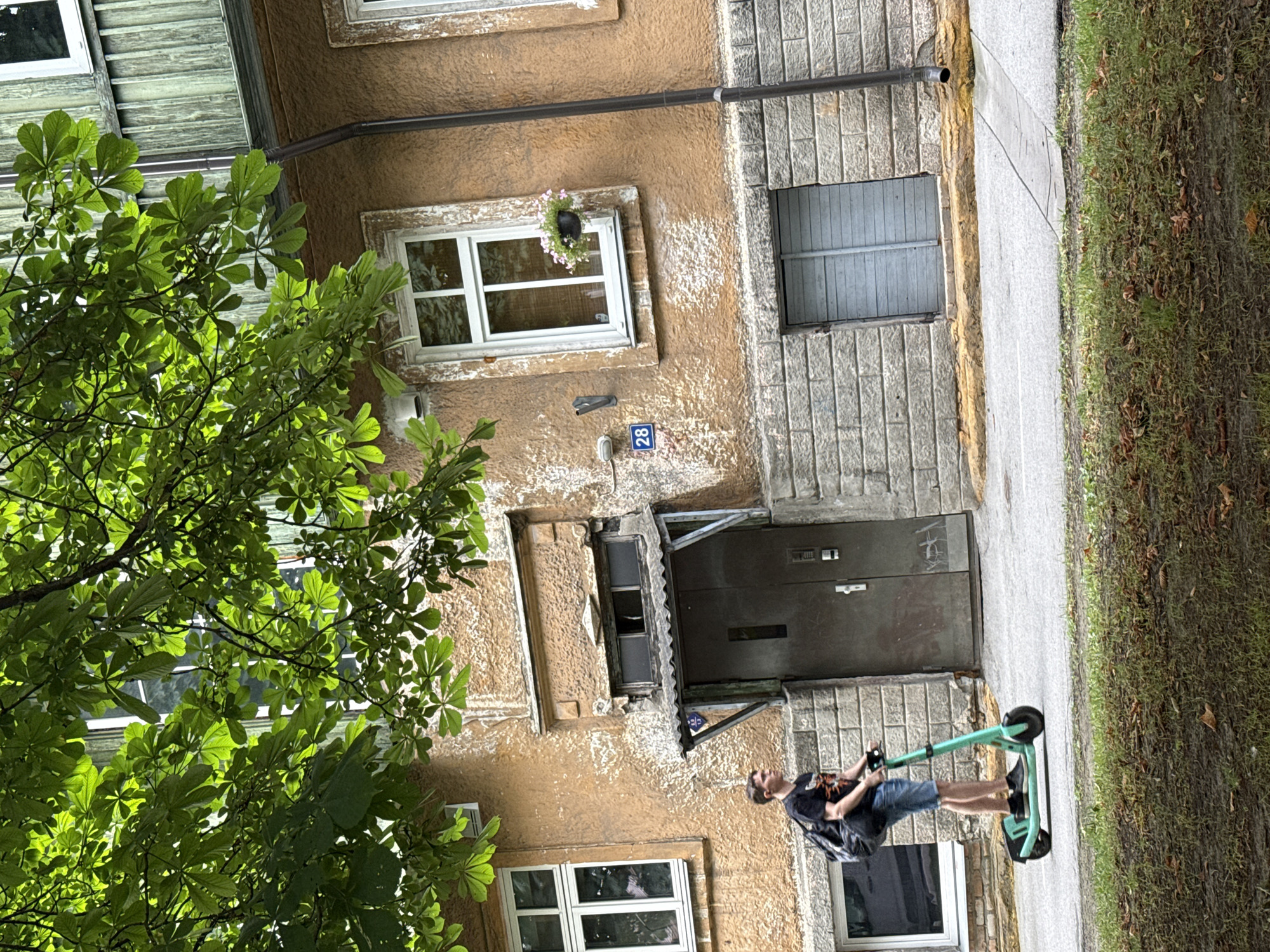 A young male, wearing a black t-shirt with an orange graphic, denim shorts, and black sneakers, is riding a teal electric scooter. He carries a black backpack. He is moving from left to right across the frame. The scooter has black handlebars and a black rear fender. The background features a weathered, orange-colored building with a green wooden upper section. The building has multiple windows, some with white frames and others with a more aged appearance. A large tree with green leaves partially obscures the top of the building. A grey, double-leafed door is visible on the building's facade, beneath a small, sloped awning. Above the door, a small, broken window is present. A blue street sign with the number "28" is affixed to the building to the left of the door. A drainpipe runs vertically down the side of the building. The ground in front of the building is paved with concrete, and a strip of grass with fallen leaves runs along the bottom of the frame. The scene is outdoors in what appears to be an urban environment.