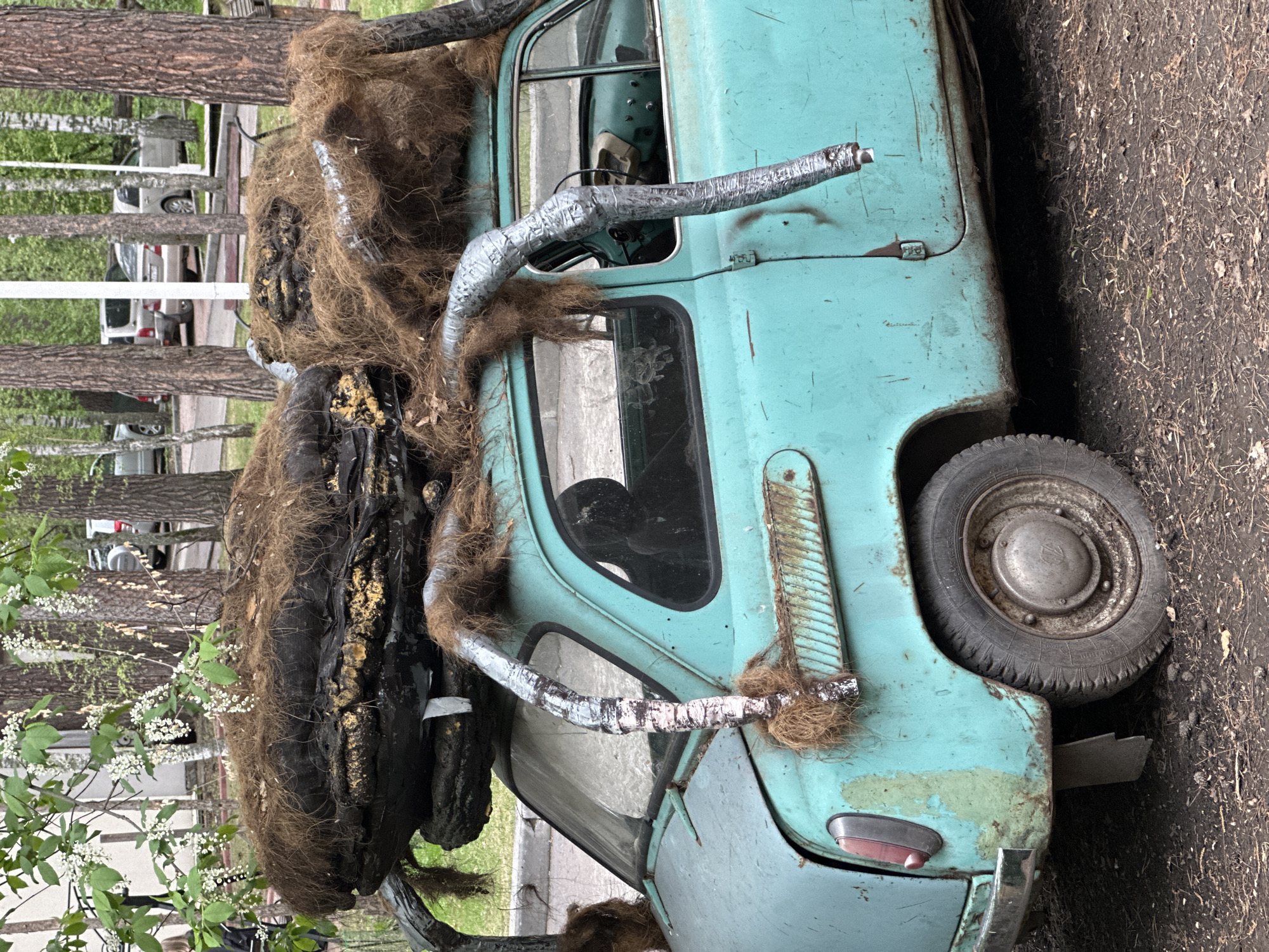 A turquoise, derelict car with a large, stylized insect sculpture attached to the roof is parked outdoors. The location is likely Berdsk municipality, Russia. The sculpture is primarily composed of dark, textured material resembling insect parts, accented with brown fibrous material and metallic-looking tubing wrapped in silver tape. The car shows signs of age and disrepair with rust and fading paint. The surrounding environment includes trees and a patch of dirt.