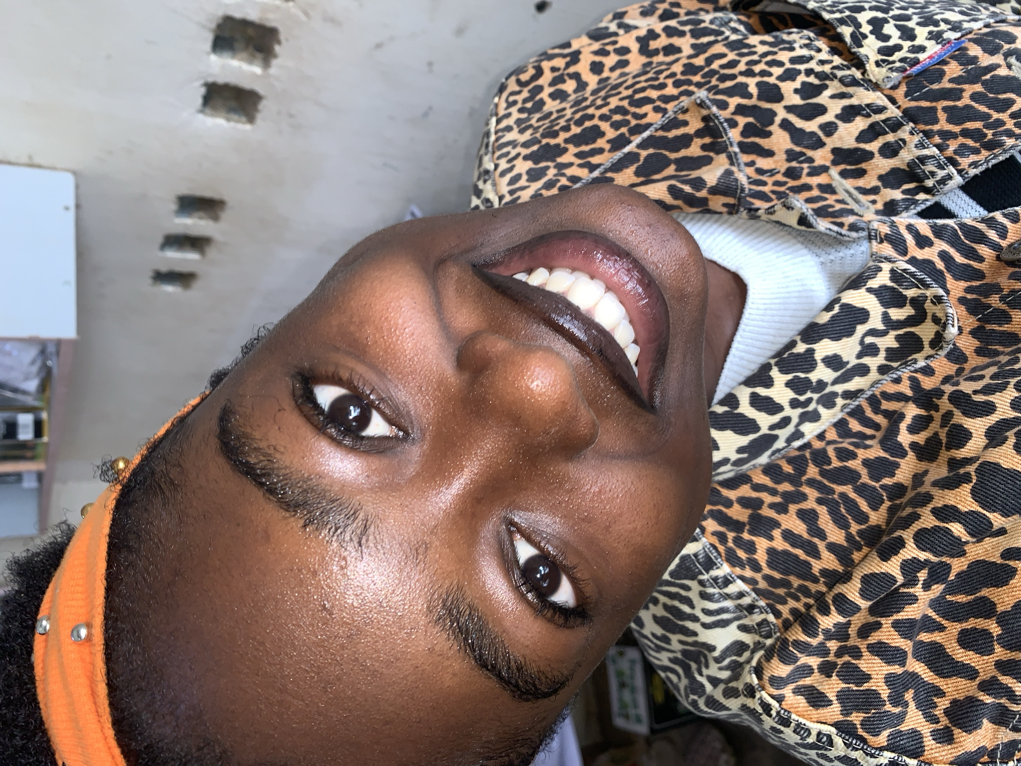 The media file is a close-up, headshot photograph of a young Black woman. She is wearing an orange headband adorned with small silver beads and a leopard-print jacket over a white turtleneck. Her eyes are open and looking at the camera, and she has a wide, bright smile, revealing her teeth. The lighting suggests it might be daytime. The background is slightly out of focus but appears to be an indoor setting with some light-colored walls and what look like some cutouts or openings in the ceiling. There is no visible text in the image. The woman's expression appears happy and confident. The setting does not provide specific cues for Jos, Nigeria, beyond the general context provided.