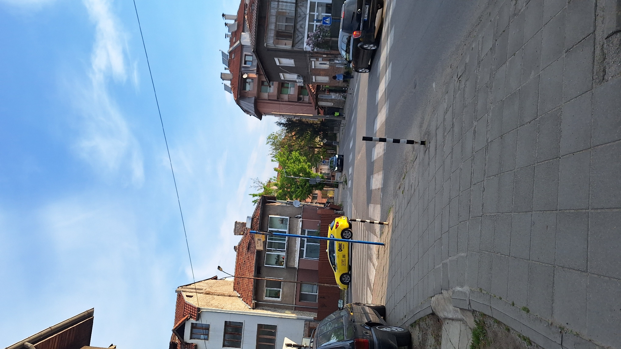 The image shows a street scene in Blagoevgrad, Bulgaria. The sky is blue with some clouds. Buildings with orange tiled roofs line the street. There are at least three cars visible, including a yellow taxi. A black and white striped pedestrian crossing is visible on the right side of the frame. A blue pole runs along the street by the taxi. At least two individuals can be seen in the distance at the pedestrian crossing.