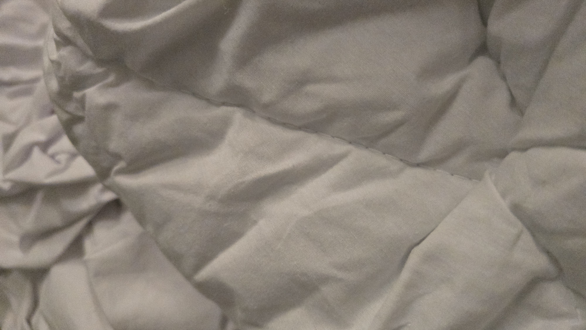 This is a close-up, abstract image of a light-colored, possibly white or off-white, quilted comforter or duvet. The fabric has a matte finish and is characterized by numerous folds and wrinkles, creating a sense of depth and texture. A prominent stitched line runs diagonally across the center of the image, forming a diamond or geometric pattern with other implied stitching. The lighting appears to be soft, casting subtle shadows within the creases of the fabric, which highlights the dimensionality of the material. There are no discernible objects, people, or environmental features outside of the textile itself. The image does not contain enough information to determine its location in Batumi, Georgia, or any other specific place.