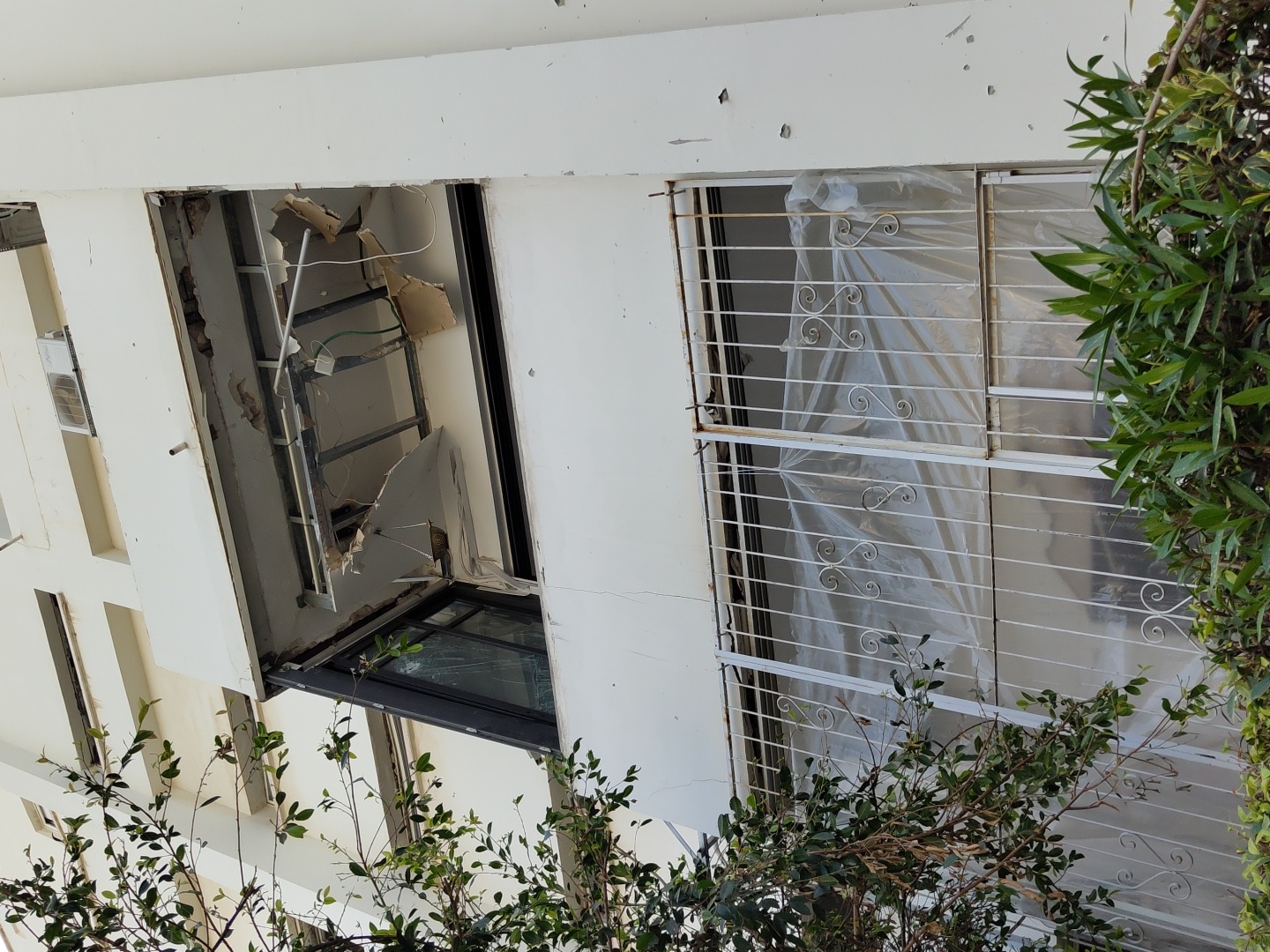 The image depicts building damage, specifically broken windows and damaged walls, in an urban setting. A portion of the wall around one window has been completely destroyed, exposing internal framework and wiring. The window itself is broken. A neighboring window has a white protective covering secured by metal bars. The exterior of the building is white. Green foliage, possibly trees or bushes, is visible in the foreground. The image is rotated. The location of Tel-Aviv, Israel is not evident in the image.