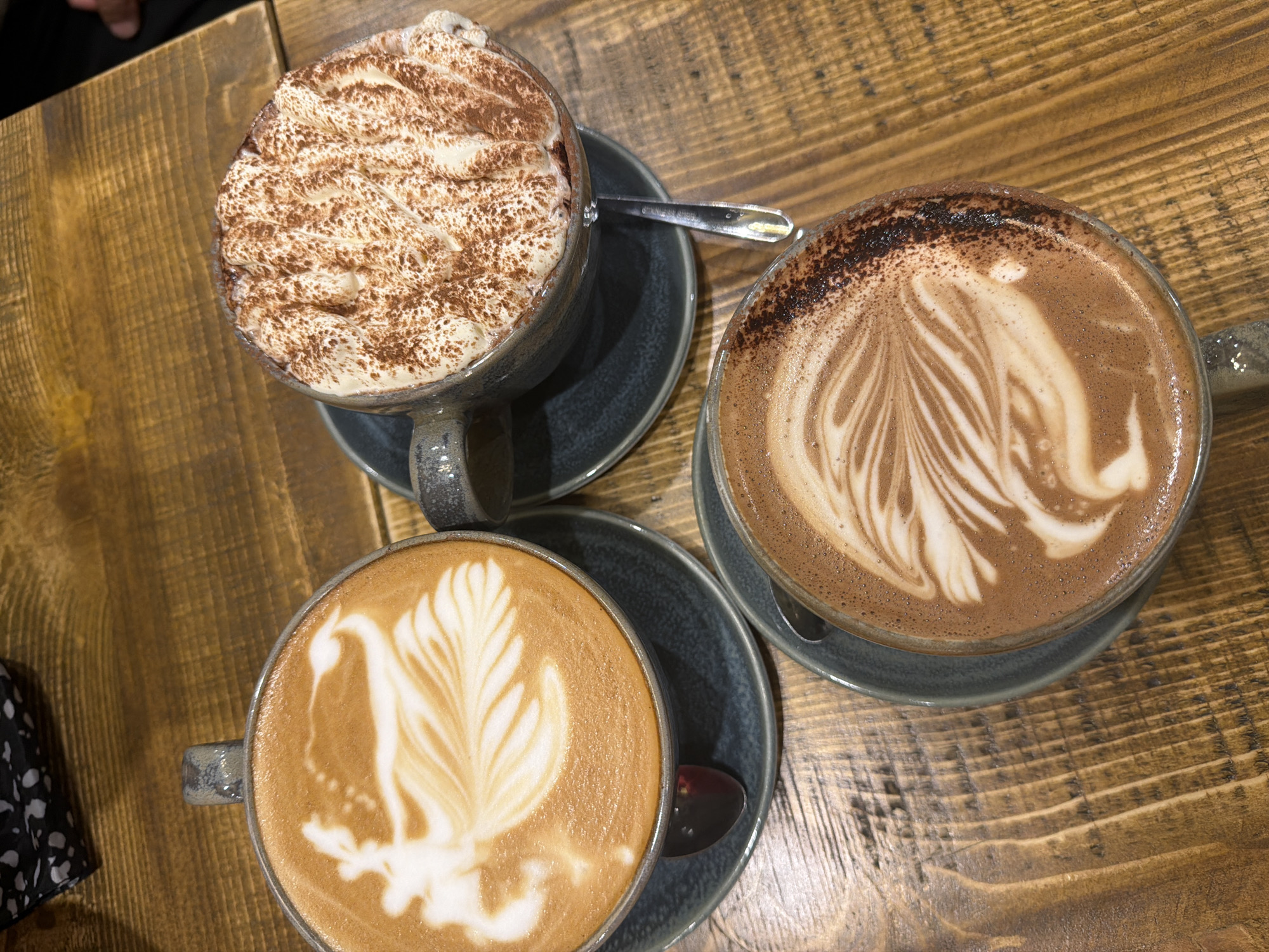 The image shows a tabletop view of three different types of beverages in mugs, likely coffee or hot chocolate, sitting on a wooden table. One mug is filled with a drink topped with a large amount of whipped cream and dusted with cocoa powder. The second mug has latte art featuring a feathered design. The third mug also has latte art. Each mug is on a saucer of the same color. A spoon rests on the saucer of the mug filled with whipped cream, and another spoon rests on the saucer of the mug with the feathered design latte art. A corner of a black and white patterned cloth is visible on the lower left. The photograph was likely taken indoors in Mole Valley, United Kingdom.