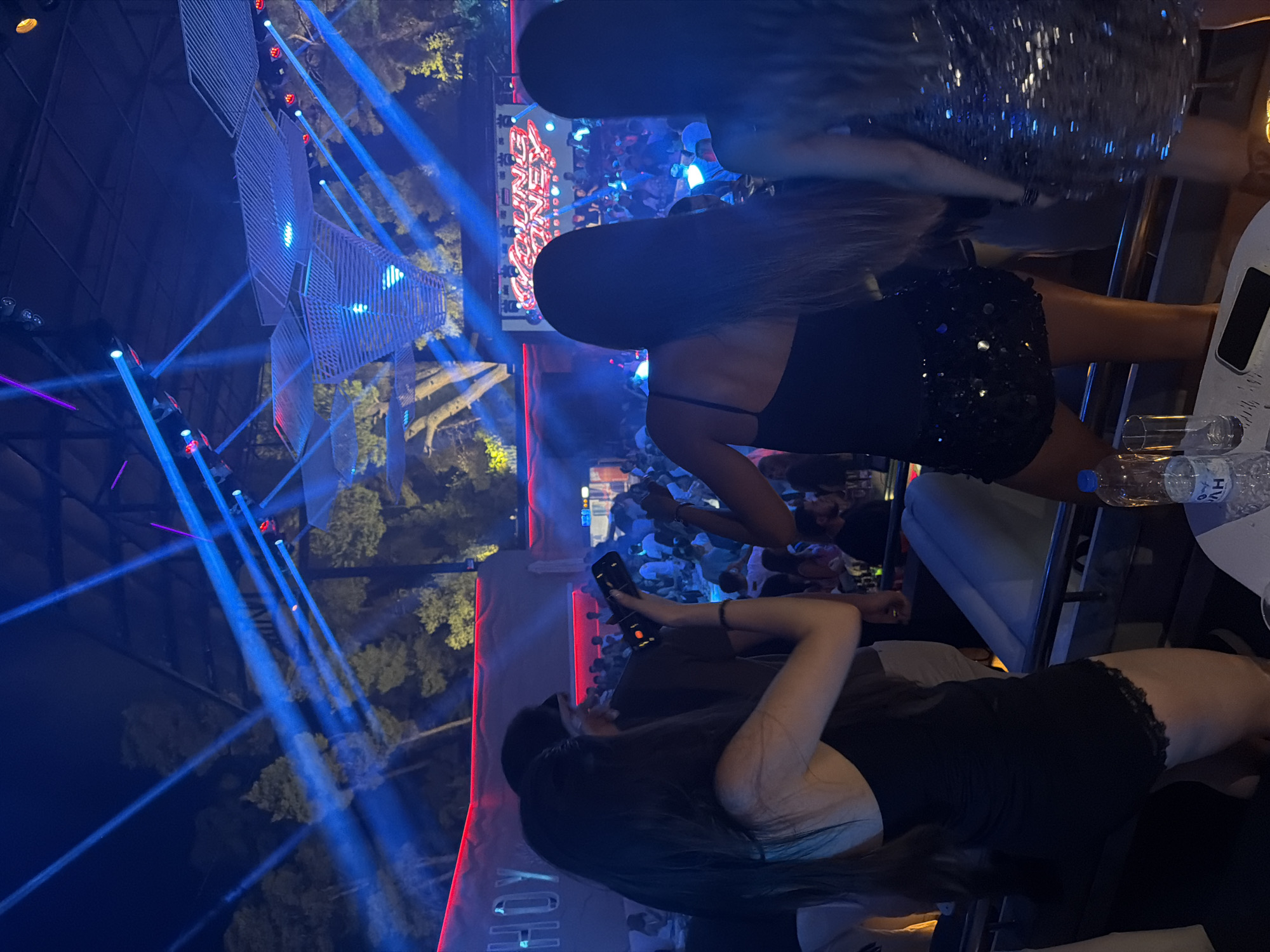 A night view of a crowded outdoor nightclub in the Kassandra Municipal Unit, Greece, features an energetic atmosphere with bright blue and purple laser lights cutting through the darkness. The foreground shows the backs of three women, two of whom are wearing short, sparkly dresses, suggesting they are enjoying a night out. One woman in the center is holding up a smartphone, possibly to record or take photos of the event. In the background, a stage with a large screen displaying "YOUNG MONEY" can be seen, indicating a performance or DJ set. The overall scene depicts a lively entertainment venue with a significant number of patrons engaged in the festivities.