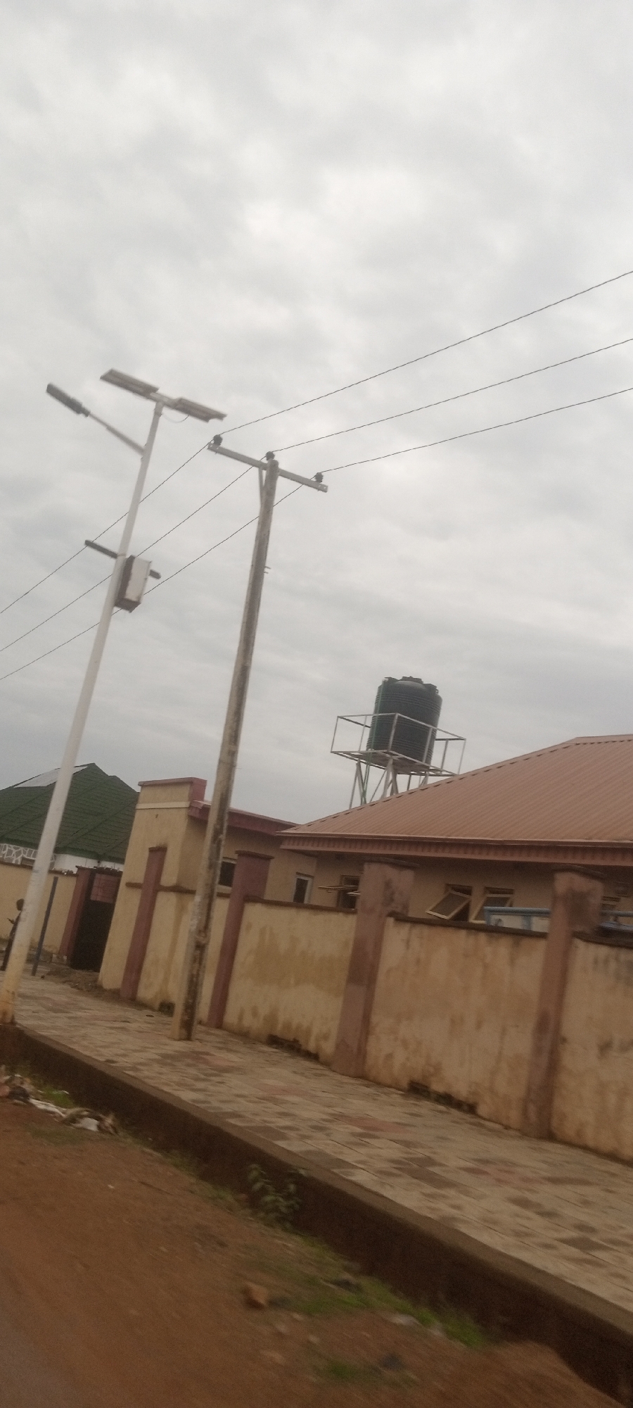 The image depicts a street scene in Narayi, Nigeria. The sky is overcast. A sidewalk lined with interlocking pavers runs alongside a building with a beige exterior and red accents. The building has a brown roof. A large black water tank sits on a metal platform atop the building's roof. An older wooden utility pole and a newer white lamp post stand along the sidewalk, and utility lines span across the sky. There are some small patches of grass and trash along the edge of the sidewalk. A person appears to be walking in the distance, next to a green roof. The perspective is from a slightly lower angle, possibly from a moving vehicle.