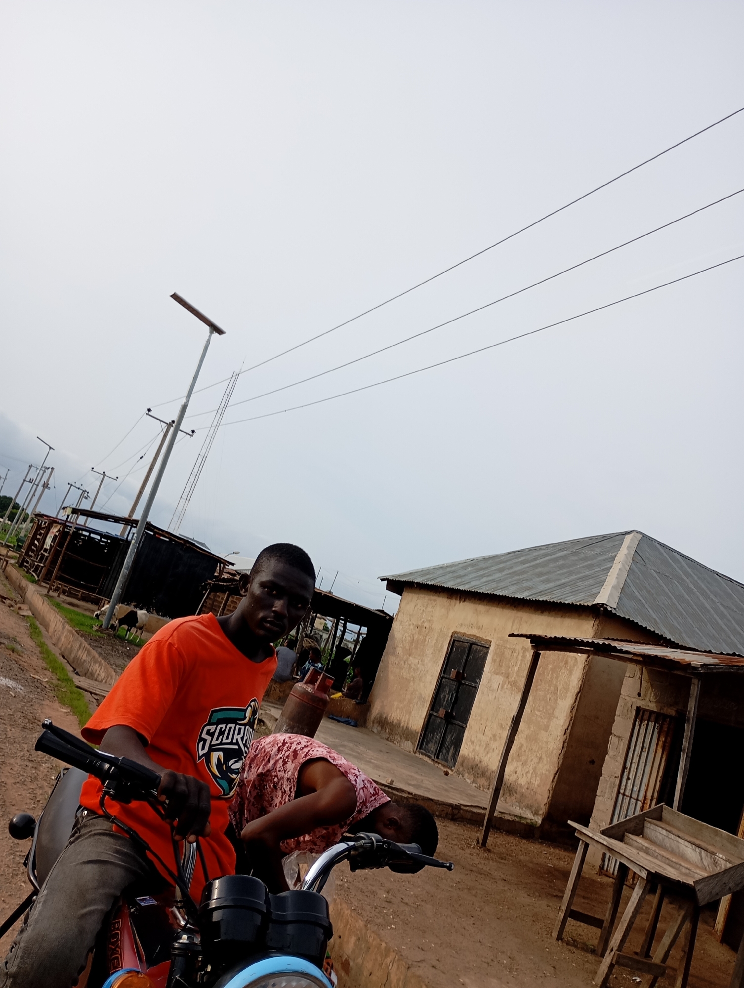 Two young men are on a red motorcycle in what appears to be a rural or semi-urban area in Shendam, Nigeria. The man in the foreground, wearing an orange t-shirt with a "Scorpio" graphic, is seated on the motorcycle, holding the handlebars. He is looking towards the camera. Behind him, another young man in a pink patterned shirt is leaning over the motorcycle, seemingly attending to it. The background features a dirt road, several simple structures with corrugated metal roofs, and utility poles with electrical wires stretching across the sky. In the middle ground, there are a few goats grazing. The overall lighting suggests an overcast day.