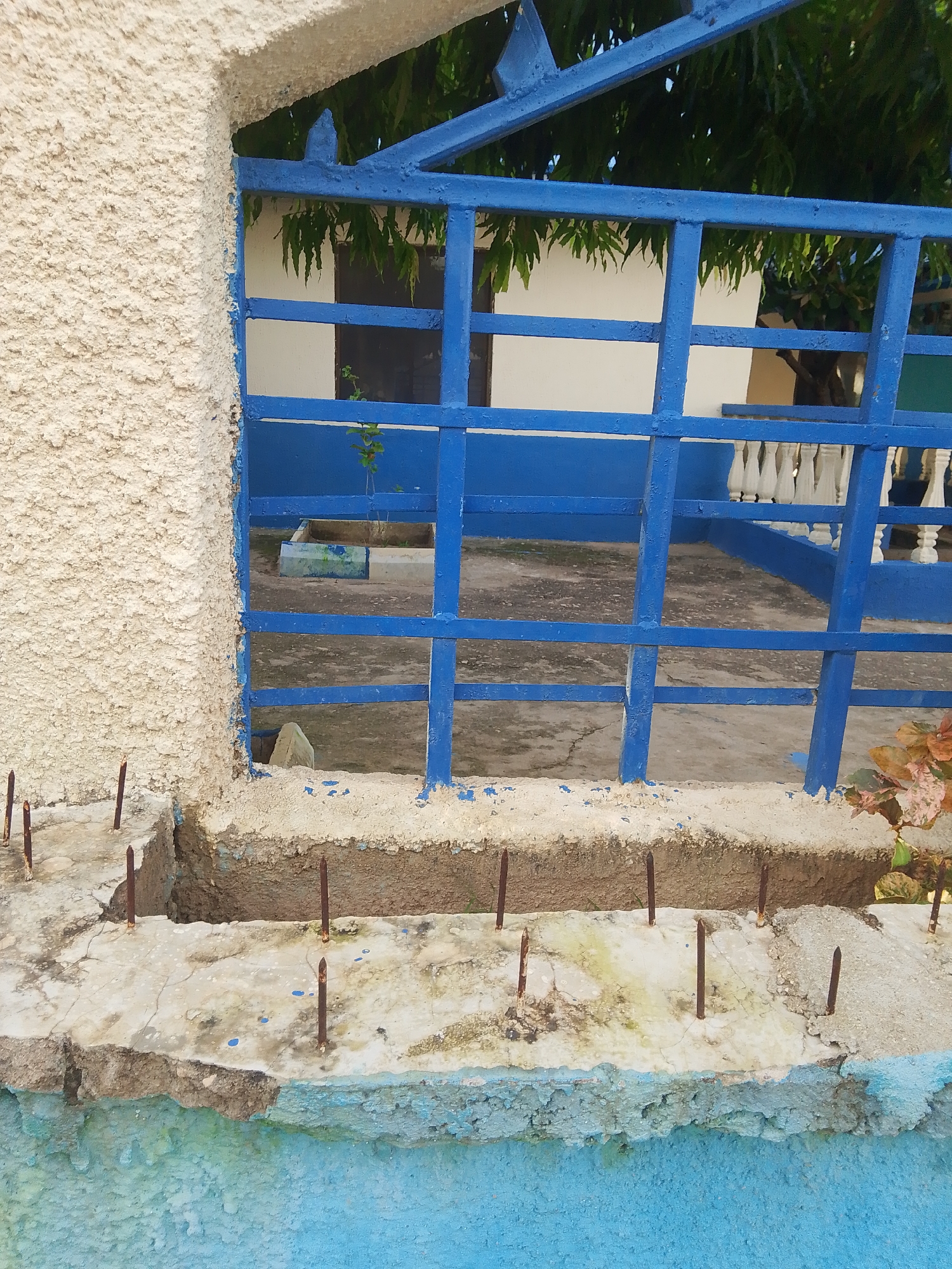 The image captures a close-up view of a damaged concrete structure with protruding rebar, likely part of a fence or boundary wall. A blue metal grate, also showing signs of wear, runs horizontally across the frame, partially obscuring the scene behind it. The concrete is rough and textured, with patches of light and dark tones suggesting weathering and possibly staining. Several rusty rebar pieces are embedded in the concrete, some pointing upwards like spikes.
Behind the blue grate, a building with white walls and a green roof is visible, along with the dense foliage of trees. A small plant is growing in a planter box. The ground appears to be a dirt or gravel surface. A white balustrade is visible in the background, suggesting a more formal or residential area beyond the immediate structure.
The overall impression is one of disrepair and neglect. The rough texture of the concrete and the exposed rebar suggest a harsh environment or a lack of maintenance. The lighting indicates it is daytime, likely sunny given the bright colors and shadows. No people are visible in the frame. The context of Jalingo, Nigeria, might imply a tropical climate, which could contribute to the weathering of the materials. There is no visible text in the image.