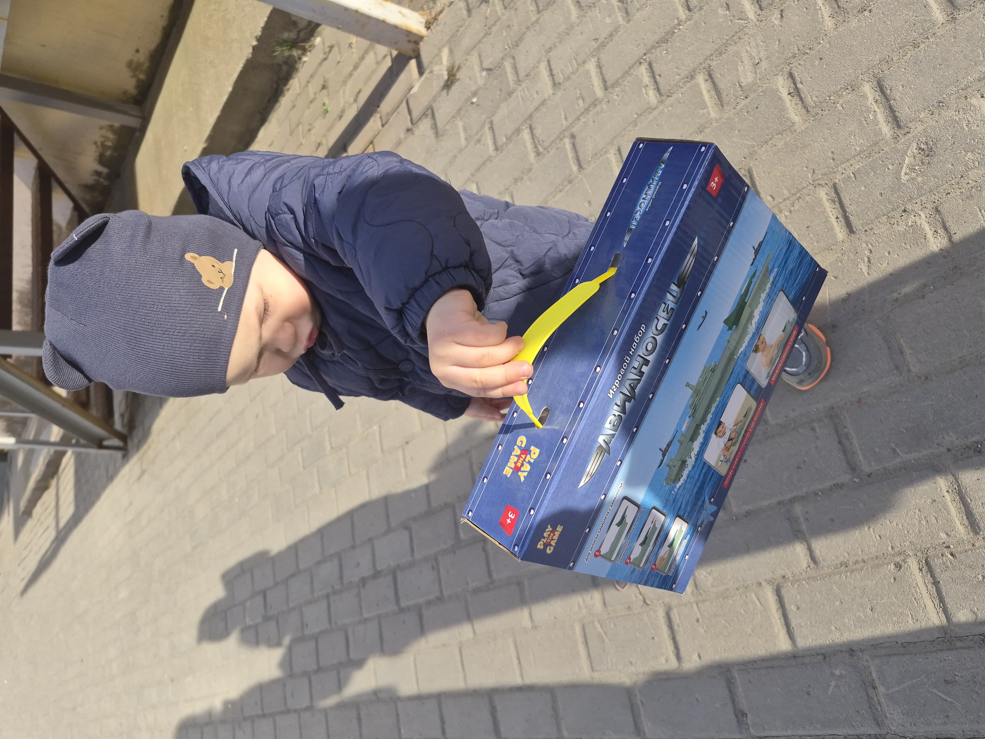 The image, taken from an overhead and slightly tilted perspective, shows a young child outdoors. The child is wearing a navy blue quilted winter jacket and a matching navy blue beanie adorned with a brown teddy bear patch. Their face is partially visible, looking down at a blue cardboard box they are holding in their hands.
The box is a toy package, prominently featuring the Russian text "Игровой набор АВИАНОСЕЦ" (Game set Aircraft Carrier), along with images of an aircraft carrier on water and airplanes. Other visible text includes "PLAY THE GAME" and "3+", indicating an age recommendation. The child is holding a bright yellow plastic piece, which appears to be inserted into a slot on the side of the box, suggesting interaction with the toy set.
The setting is an outdoor paved area, likely a sidewalk or courtyard, composed of light-colored rectangular paving stones. Strong sunlight casts distinct shadows on the ground, indicating daytime. In the background, a metal structure, possibly part of a railing or stairs, is visible. The scene captures a moment of the child engaged with their new toy on what appears to be a cool, sunny day.