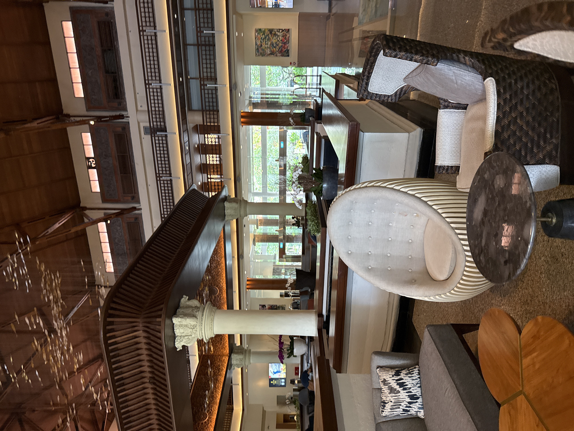 This image shows the interior of a building, likely a hotel lobby, located in Nusa Dua, Indonesia. The scene includes various pieces of furniture such as armchairs, sofas, and tables arranged in a seating area. Architectural features are prominent, including a multi-story interior design with decorative columns, ornate detailing on the staircases, and window-lined walls. There is ambient light filtering through the windows and from overhead fixtures. The furniture is placed on a carpeted floor. The scene is devoid of human presence.
