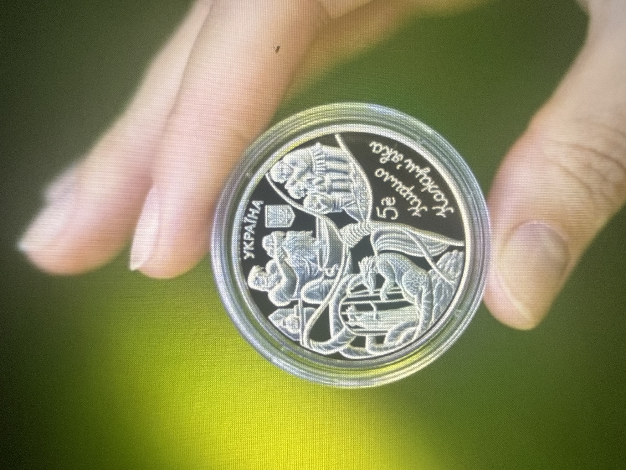 The image shows a person's hand holding a coin with a silver rim. The coin has a black background with engraved silver imagery. At the top of the coin, the word "УКРАЇНА" (Ukraine) is visible, along with a crest. The central image appears to depict figures engaged in some activity, possibly related to pottery or craftsmanship, with a coiled snake-like creature at the bottom. Below the imagery, the number "5" is present, followed by Cyrillic text that reads "Кирило Кожум'яка" (Kyrylo Kozhumiaka). The background of the image is a blurred green, suggesting an outdoor setting. There is no identifiable information within the image that places the scene specifically in the city of Dykivka, Ukraine.