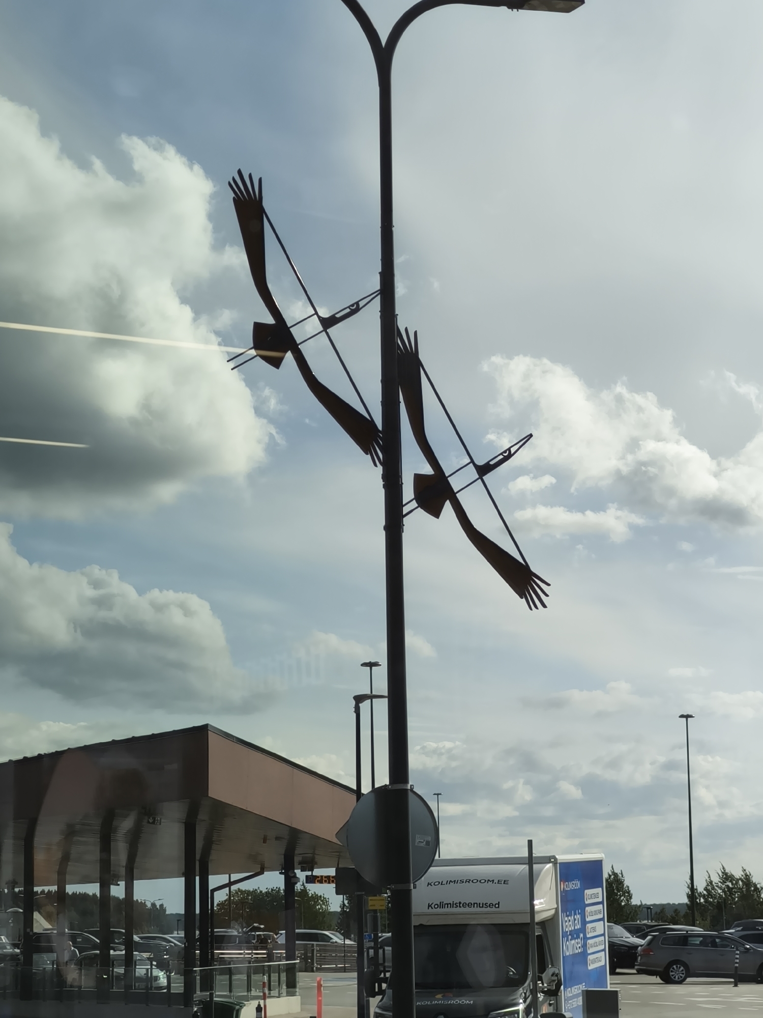 This image was taken in Tallinn, Estonia, likely at a parking lot or toll booth area, given the presence of a covered structure with supporting pillars and a road with vehicles. Dominating the sky is a tall lamppost with a sculpture of three birds in flight attached to it. The birds are depicted as metallic silhouettes with outstretched wings and open hands instead of feet. The sky is partly cloudy with a mix of sunlight and shadow. In the foreground, a white truck with blue lettering is visible, indicating a service related to moving or storage, with "KOLIMISROOM.EE" and "Kolimisteenused" printed on its side. Other vehicles are parked in the background.